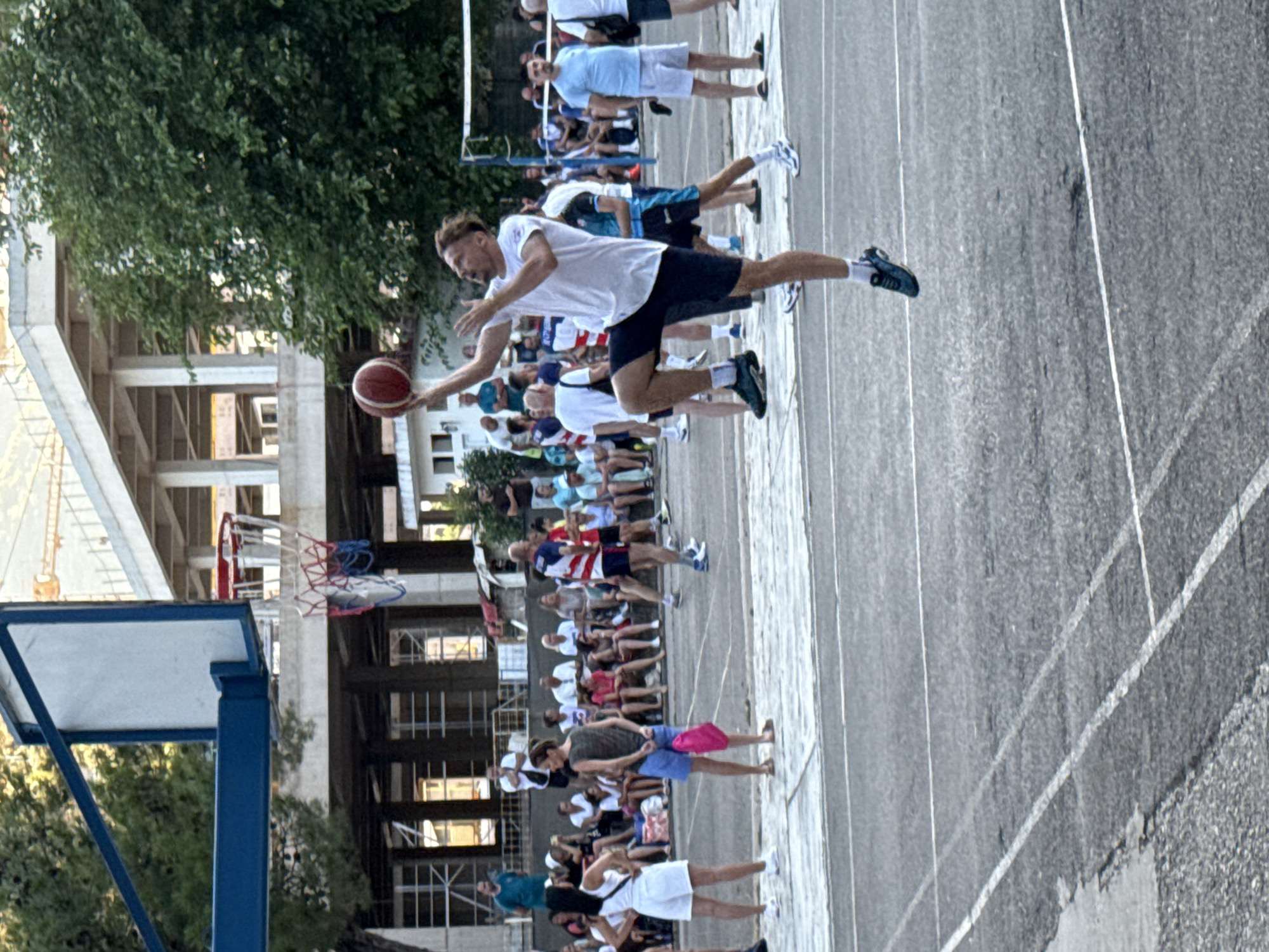 In Budva, Montenegro, a man in mid-air is attempting a basketball shot on an outdoor court. He wears a white t-shirt and dark shorts, with his right arm extended and holding the basketball. His left arm is bent at the elbow. The court is paved with asphalt and marked with white lines. In the background, spectators are gathered, some seated and others standing, observing the game. Behind the spectators, there are trees and buildings under construction, suggesting an urban environment. A basketball hoop with a net is visible.