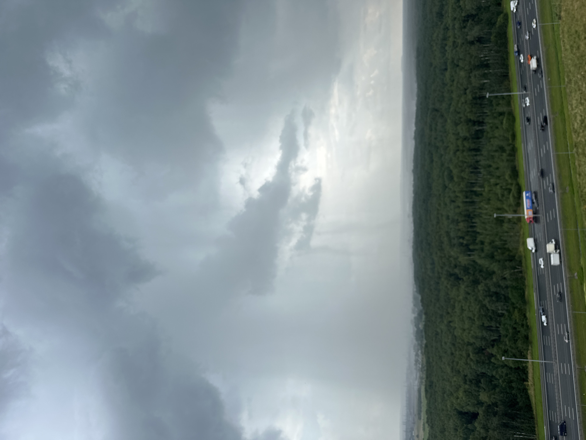 A wide-angle, high-angle shot captures a stormy sky over a vast forest and highway in Severnaya Samarka, Russia. Heavy, dark gray clouds dominate the upper portion of the frame, with shafts of light breaking through, creating a dramatic effect. A dense, dark green forest stretches across the middle ground, with a multi-lane highway running in front of it. Numerous vehicles, including cars and trucks, are visible on the wet highway, suggesting recent or ongoing rain. The overall atmosphere is one of inclement weather, with a sense of vastness and the power of nature.