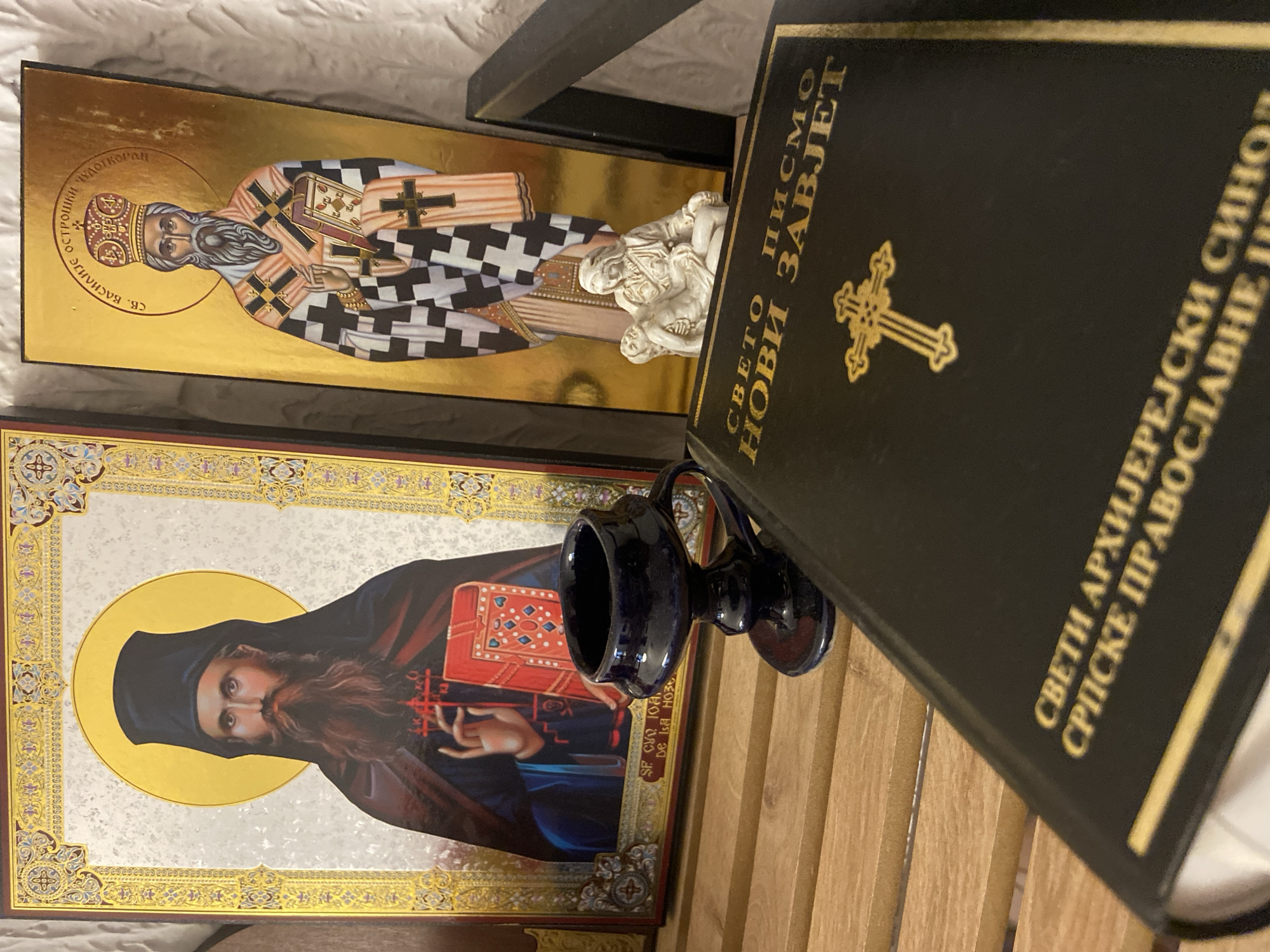 An indoor scene in Sutomore, Montenegro, displays a static arrangement of religious items on a light-colored wooden slatted surface, set against a light, textured background. Two prominent Orthodox icons stand upright. The left icon, framed in gold with ornate patterns, depicts a bearded male saint identified as Saint John of the Ladder, dressed in dark monastic robes, with a golden halo, raising his right hand in blessing and holding a closed red book in his left. The right icon, primarily gold-toned, portrays a bearded male saint identified as Saint Basil of Ostrog the Wonderworker, wearing a bishop's mitre and black-and-white checkered vestments, with a golden halo, presenting an open book with his left hand.
In the middle ground, a small white Pieta figurine, depicting a robed woman cradling a deceased man, is positioned in front of the lower section of the right icon. A dark blue, ceramic, goblet-shaped object with a handle rests on its pedestal, positioned to the left of the Pieta figurine and in front of the left icon. In the immediate foreground, a large, black-bound book with gold lettering and a prominent gold cross emblem lies angled. The Cyrillic text on its cover reads "СВЕТО ПИСМО НОВИ ЗАВЈЕТ" (Holy Scripture New Testament) and further specifies "СВЕТИ АРХИЈЕРЕЈСКИ СИНОД СРПСКЕ ПРАВОСЛАВНЕ ЦРКВЕ" (Holy Episcopal Synod of the Serbian Orthodox Church). No active persons are visible.