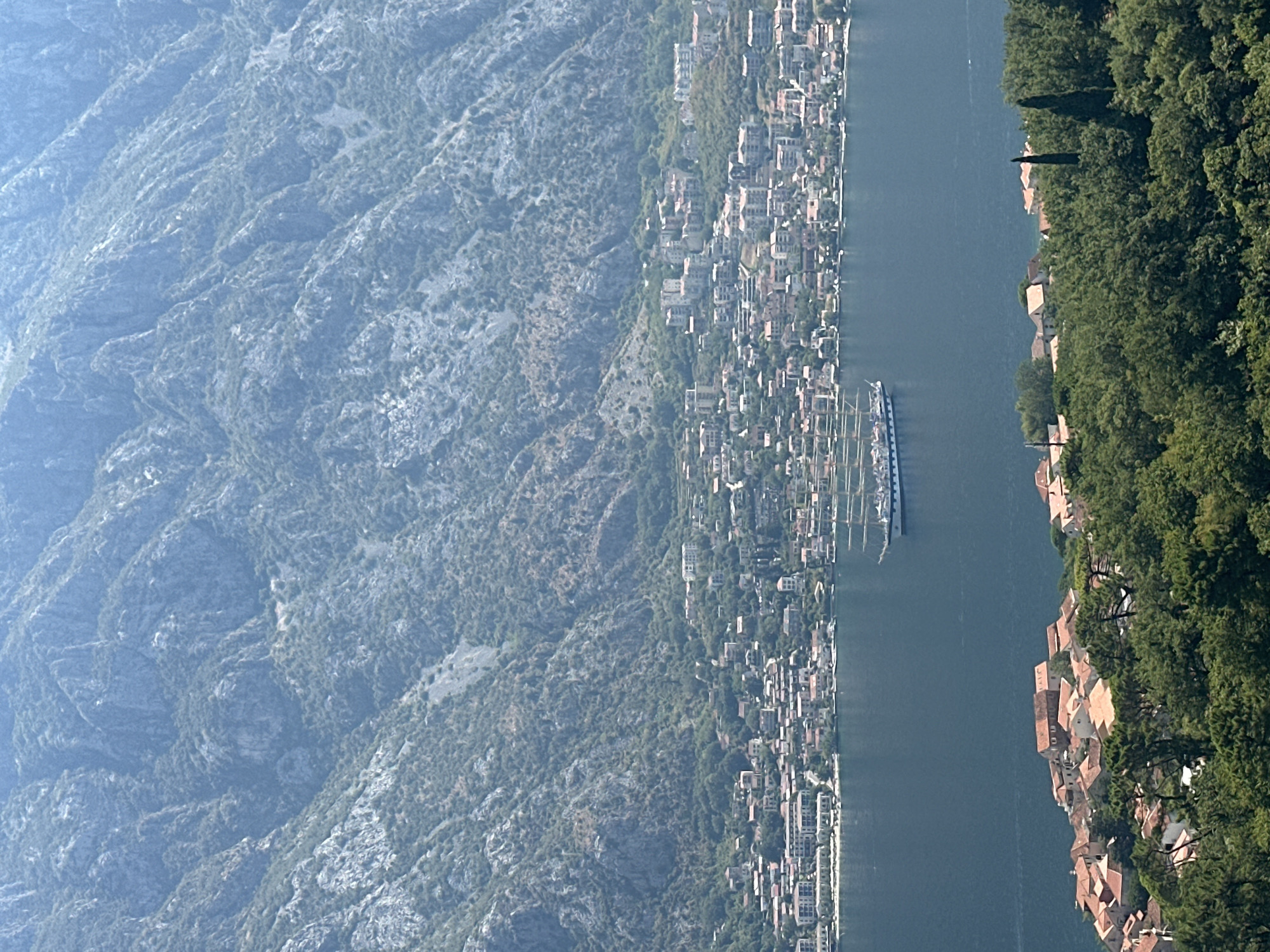 The image is an aerial view of the town of Prčanj, Montenegro, situated along a bay with a large mountain range in the background. The foreground features a dense cluster of green trees and red-roofed buildings. In the middle ground, a wide expanse of dark blue water is visible, with a large, multi-masted sailing ship positioned in the bay. Beyond the water, a coastal town with numerous buildings, many with red roofs, stretches along the shoreline, backed by a steep, rocky mountain covered in sparse vegetation. The sky is a pale blue, indicating a bright, sunny day.