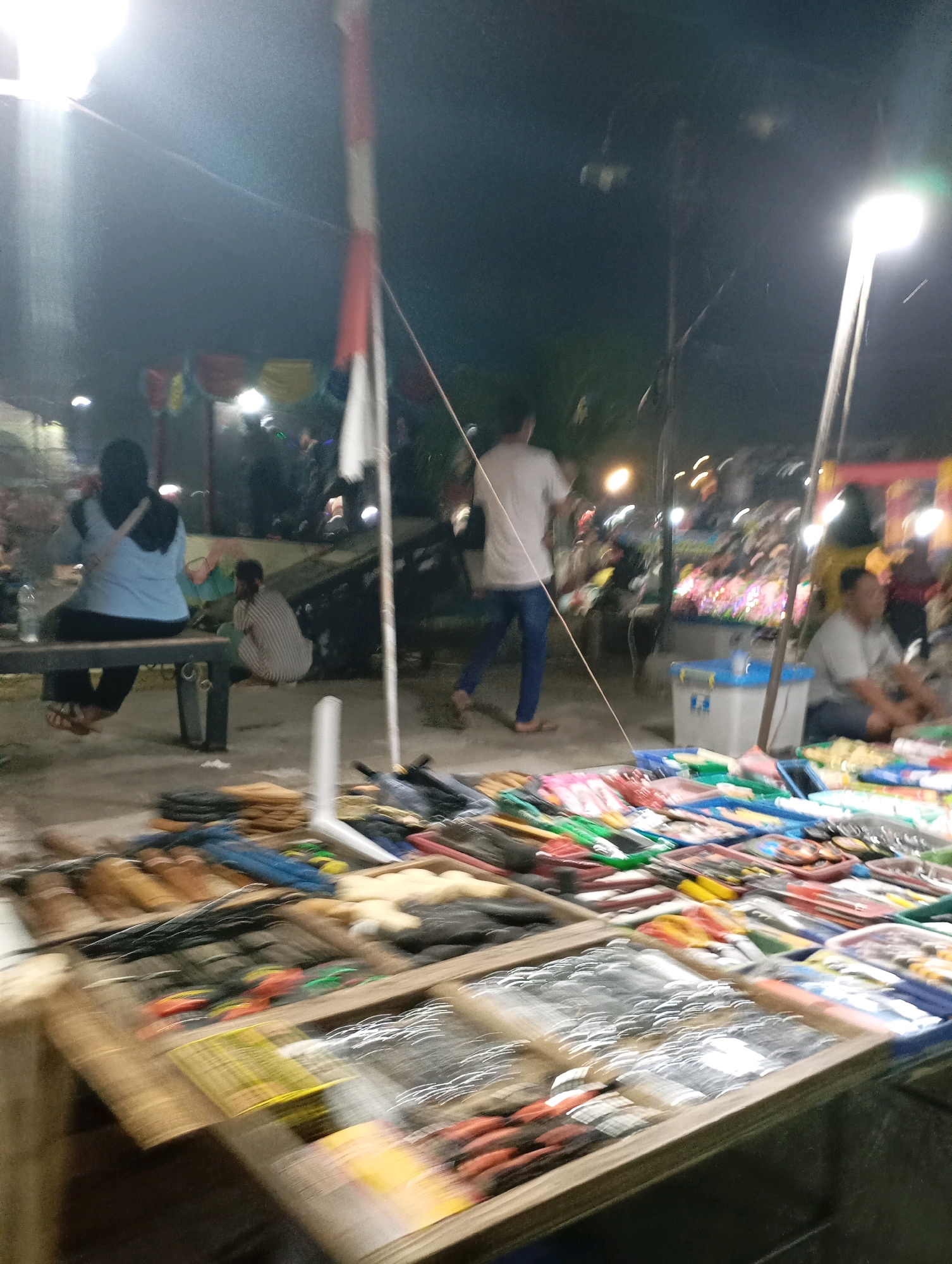 This is a nighttime photograph taken at a market stall in Kecamatan Haurgeulis, Indonesia. In the foreground, a wooden stall displays various items, including what appear to be fishing lures or small tools, packaged in plastic. The items are arranged in trays and are blurred due to motion. Behind the stall, several people are visible. On the left, a person wearing a blue shirt and a dark headscarf is seated on a bench. Next to them, another person in a striped shirt is crouched down. Further back, a person in a white t-shirt and blue jeans walks away from the camera. In the background, market stalls are set up with colorful lights and various goods. A flagpole with a red and white flag is also present, along with streetlights illuminating the scene. The ground appears to be paved.