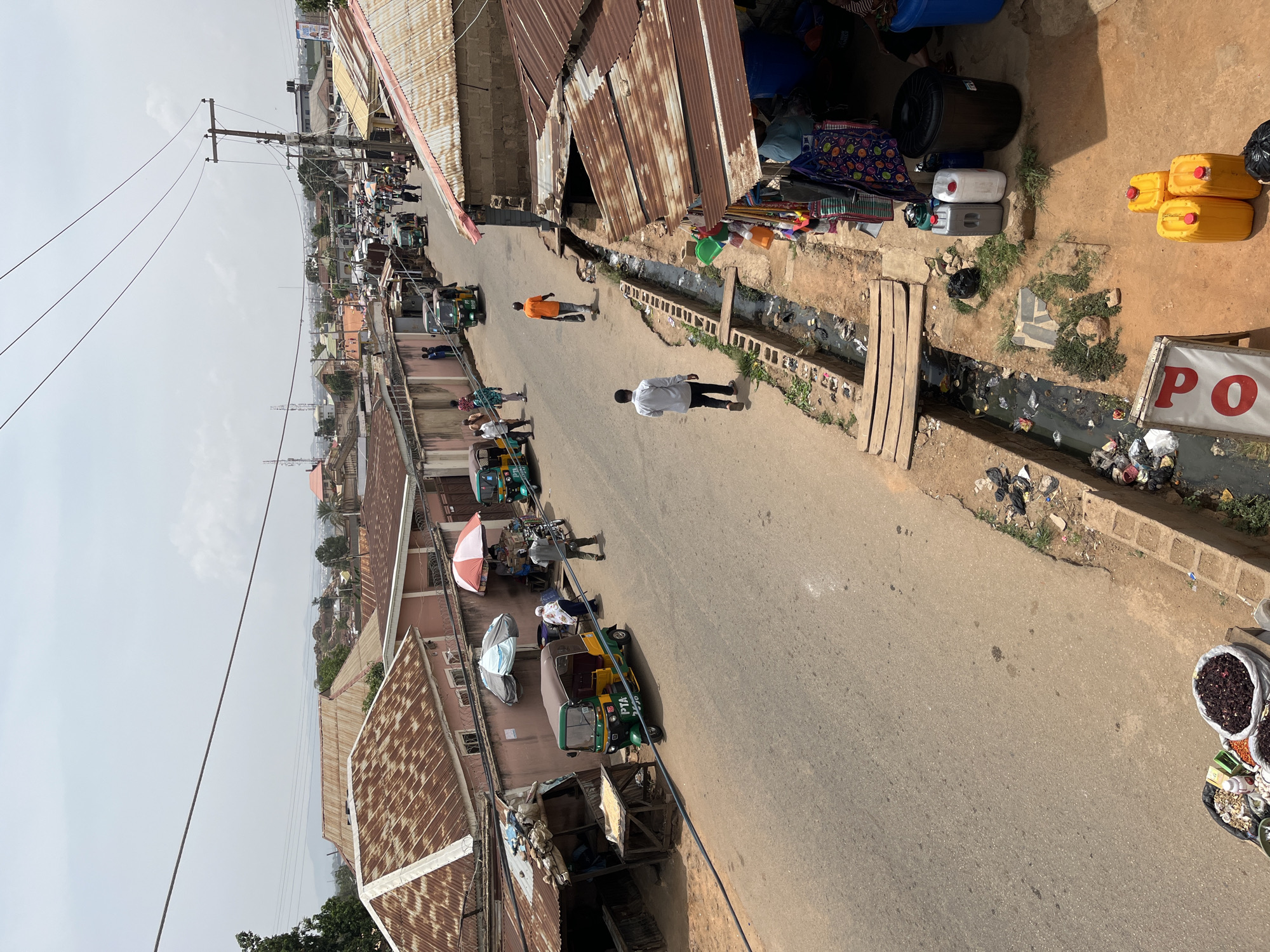 The image depicts a street scene in Gura Top, Nigeria. The street is lined with buildings, some with corrugated metal roofs. A drainage ditch runs alongside the road, filled with trash. Several people are walking along the road. There are also motorized tricycles (tuk-tuks) parked and in use. Items for sale are displayed on the side of the road. Power lines stretch across the sky above the street.