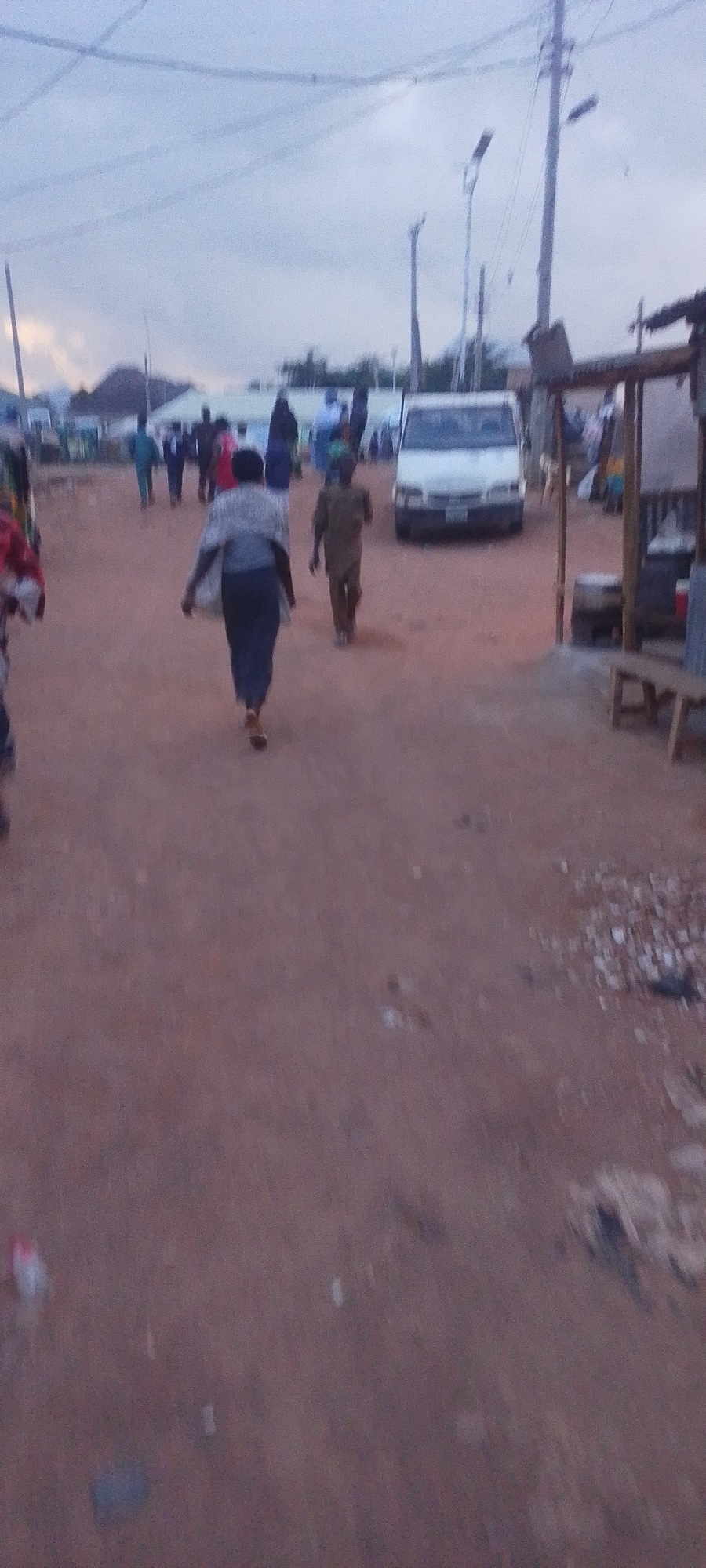 The image depicts a street scene in Gura Top, Nigeria, under an overcast sky. The ground is a reddish-brown dirt. In the foreground, two figures are walking away from the camera. The person on the left is wearing a dark skirt and a light-colored shawl or wrap over a grey top, with brown shoes. The person on the right is wearing a tan-colored outfit, possibly a kaftan, with tan shoes. In the middle ground, a white van is parked. Further in the background, a line of people are visible, some standing and others walking. Buildings with visible rooftops and a tall utility pole with streetlights are also present. A small market stall with a bench and some items on display is visible on the right side of the image. The overall impression is that of a bustling, informal urban or semi-urban environment.
