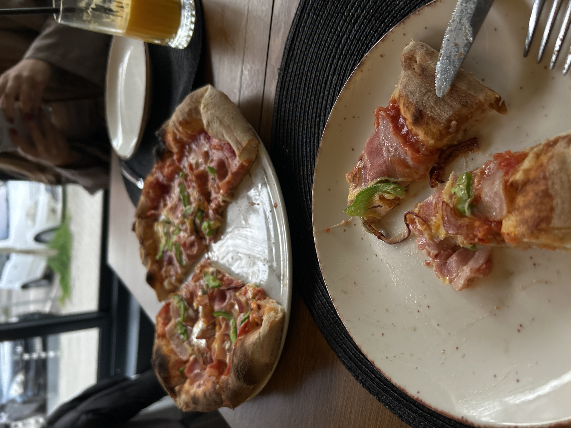 The image captures a close-up, indoor scene in what appears to be a restaurant setting. Two plates of pizza, each containing cut slices, dominate the frame. The pizza features a crust with toppings, seemingly ham, tomato sauce, and green peppers. On the right plate, a knife and fork rest near the slices, suggesting a recent interaction with the food. To the left, a partially visible glass contains an orange beverage, possibly juice. The background reveals a window with a view of what seems to be a street scene, while parts of a person's arm and clothing are visible, indicating a human presence. The location is likely in Sofia, Bulgaria, but this cannot be confirmed by the image alone.