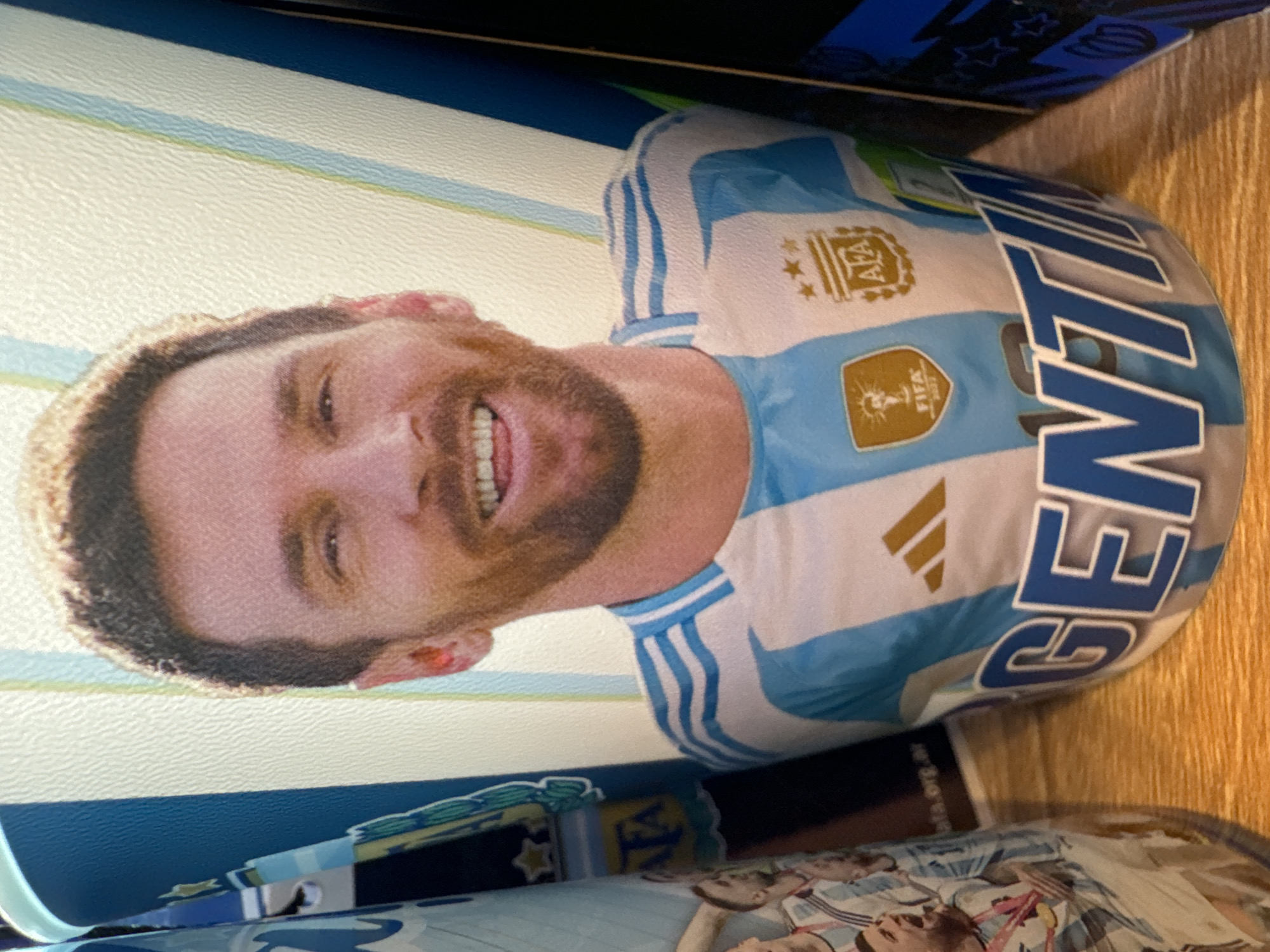 The image shows a close-up of a water bottle with a picture of Lionel Messi wearing an Argentina national team jersey. The background is a blurred tabletop with other objects partially visible. The water bottle is white with light blue stripes and features the Argentina Football Association (AFA) logo, FIFA World Cup logo, and Adidas logo, along with the word "Argentina." The picture on the bottle is a portrait of Messi, smiling and facing slightly to the left. The surrounding environment appears to be a room setting. Buenos Aires is not directly represented in the image, but the Argentina jersey alludes to the country and its football team.