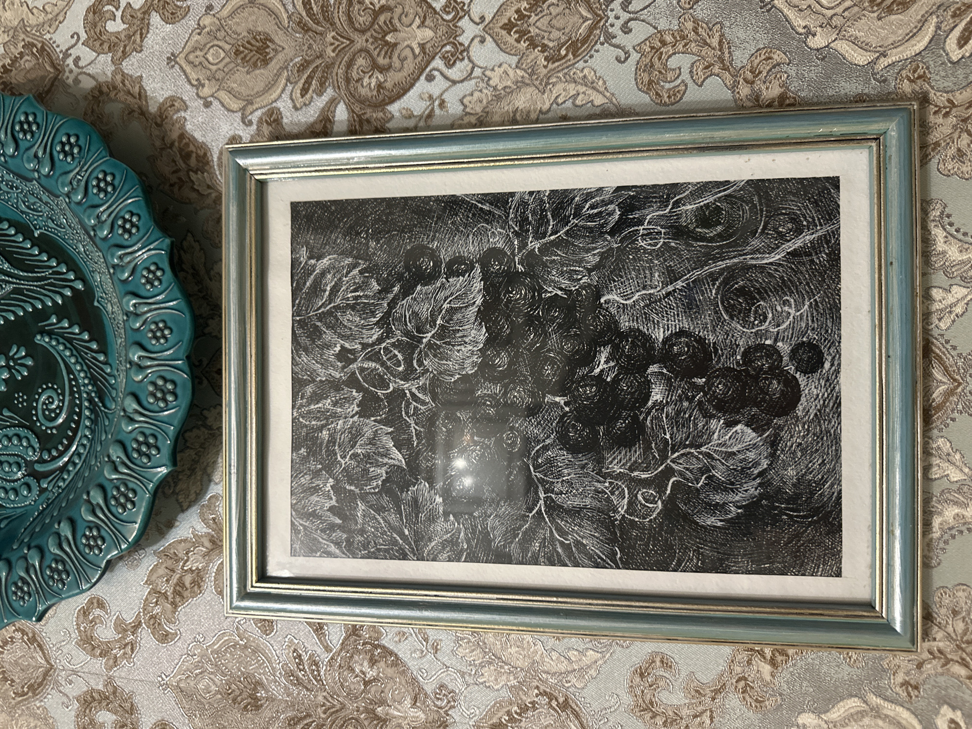 A framed black and white drawing of grapes and leaves hangs on a wall. Above the drawing is a teal decorative plate with embossed floral designs. The wall has a patterned wallpaper with tan and light blue damask designs. The location is Unknown, Unknown.