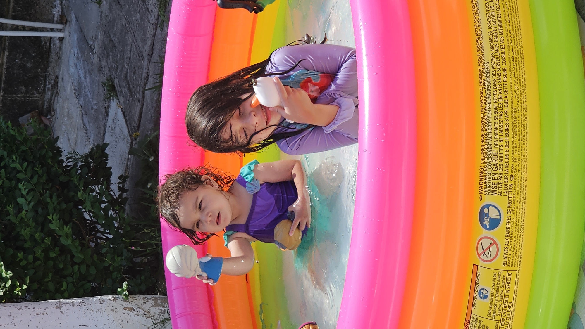 Two young girls are playing in a colorful inflatable pool outdoors. The older girl is wearing a purple long-sleeve shirt and holding a white water gun, while the younger girl is wearing a purple dress with blue accents and holding a yellow toy. The pool has rainbow-colored stripes. In the background, there is a concrete wall, green plants, and a metal structure. The location is San Bernardino Tlaxcalancingo, Mexico.