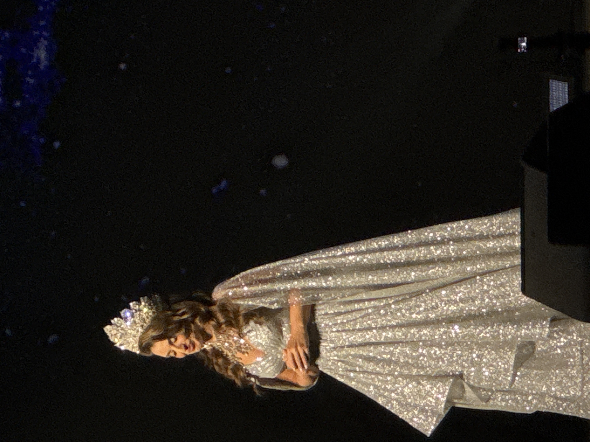 A woman wearing a sparkling gown and a crown is seen in the city of Unknown, Unknown. She is adorned with jewelry. The background is dark with scattered light dots. A dark structure is visible in the upper right corner of the frame.