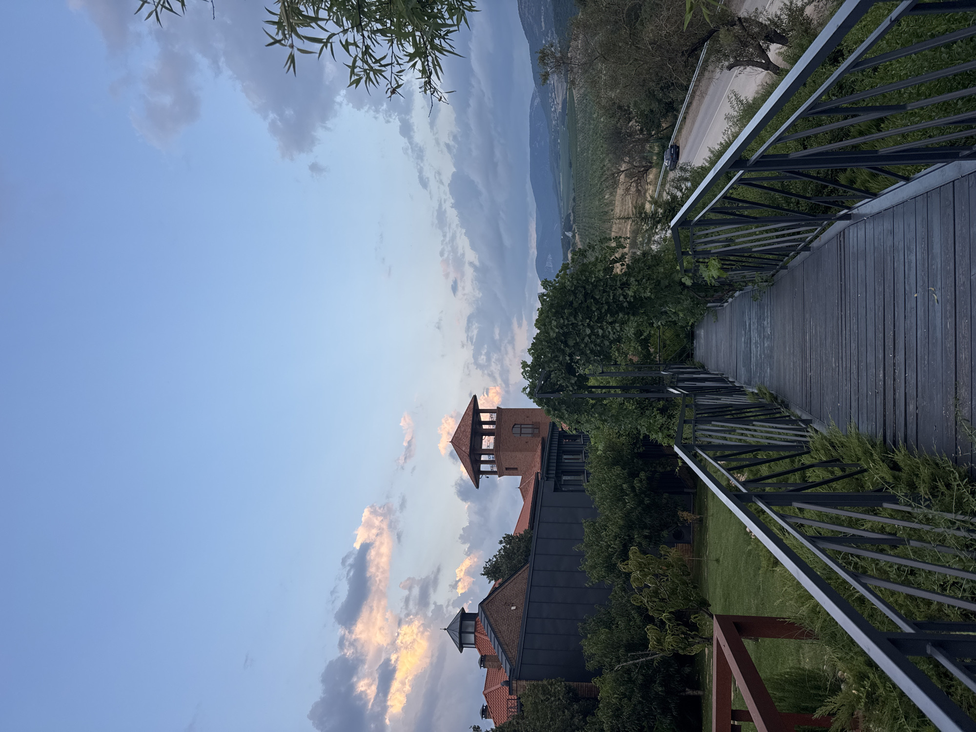 The image captures a scenic view from an elevated wooden walkway with metal railings, leading towards a landscape in Balaklavsky District, Ukraine. The walkway overlooks a lush green area with trees and a building complex featuring red-tiled roofs and a brick tower. In the background, rolling hills and a road with a single car are visible under a partially cloudy sky with soft, warm light from the setting sun. The overall atmosphere is tranquil and picturesque.