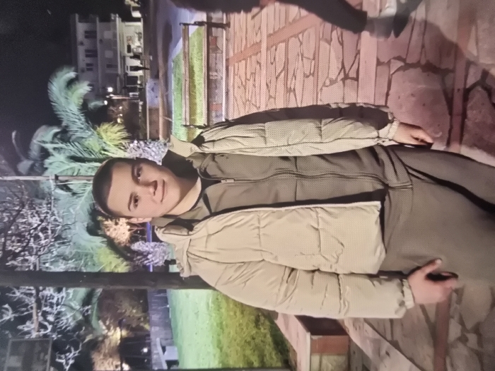 A young male stands centered in the frame, outdoors at night in a public area within the city of Samokov, Bulgaria. He is a light-skinned individual, appearing to be in his late teens or early twenties, with a closely cropped haircut. He wears a beige puffer jacket over a dark olive-green zippered track jacket and matching track pants. His right hand is positioned at his right side. He looks directly at the camera with a neutral expression.
The background consists of a park or square environment. To the left and behind the subject, there are several tall palm trees with visible fronds and green grass. A light-colored stone bench is partially visible behind the subject on the left. In the mid-ground, a decorative wall or planter box with green elements runs horizontally. In the far background, a multi-story building with illuminated windows is visible. To the right, a second individual is partially visible from the waist down, wearing dark pants and light-colored shoes, appearing to move away from the camera. The ground is paved with irregular stone slabs. The scene is illuminated by artificial light sources from streetlights or park lighting, indicating nighttime conditions.