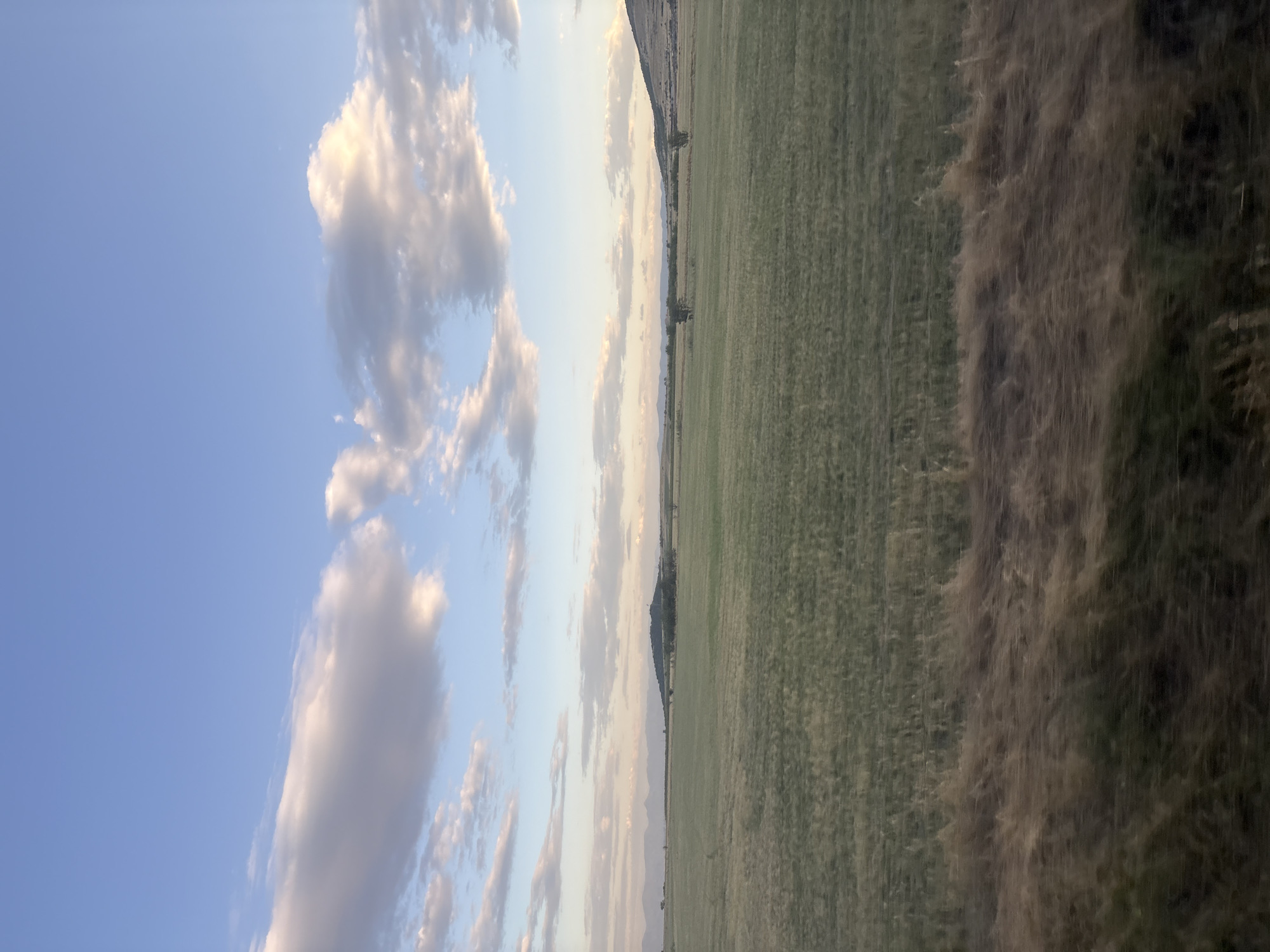 The image depicts a vast, open field under a sky with scattered clouds. The field is covered in green grass, with a strip of dry, brown grass in the foreground. In the distance, rolling hills and mountains are visible. The scene appears to be in a rural area, possibly in or near Naiden Gerovo, Bulgaria, given the natural landscape. There are no people or man-made structures immediately apparent in the image. The lighting suggests it might be late afternoon or early evening, with soft sunlight illuminating the clouds.