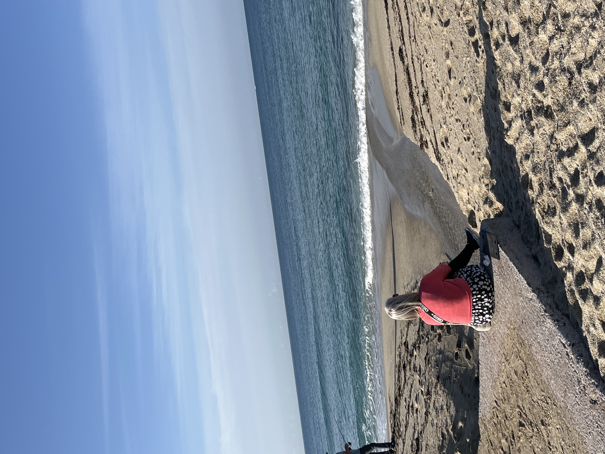 A woman is sitting on a sandy beach along the coast of Alanya, Turkey, near the Mediterranean Sea. The sky is mostly clear with a few streaks of clouds. The ocean waves break on the shore. A man is fishing in the water at the lower left corner of the frame. The woman is fair-skinned and has blonde hair. She is wearing a long-sleeved red shirt, and spotted pants. She appears to be wearing black shoes.