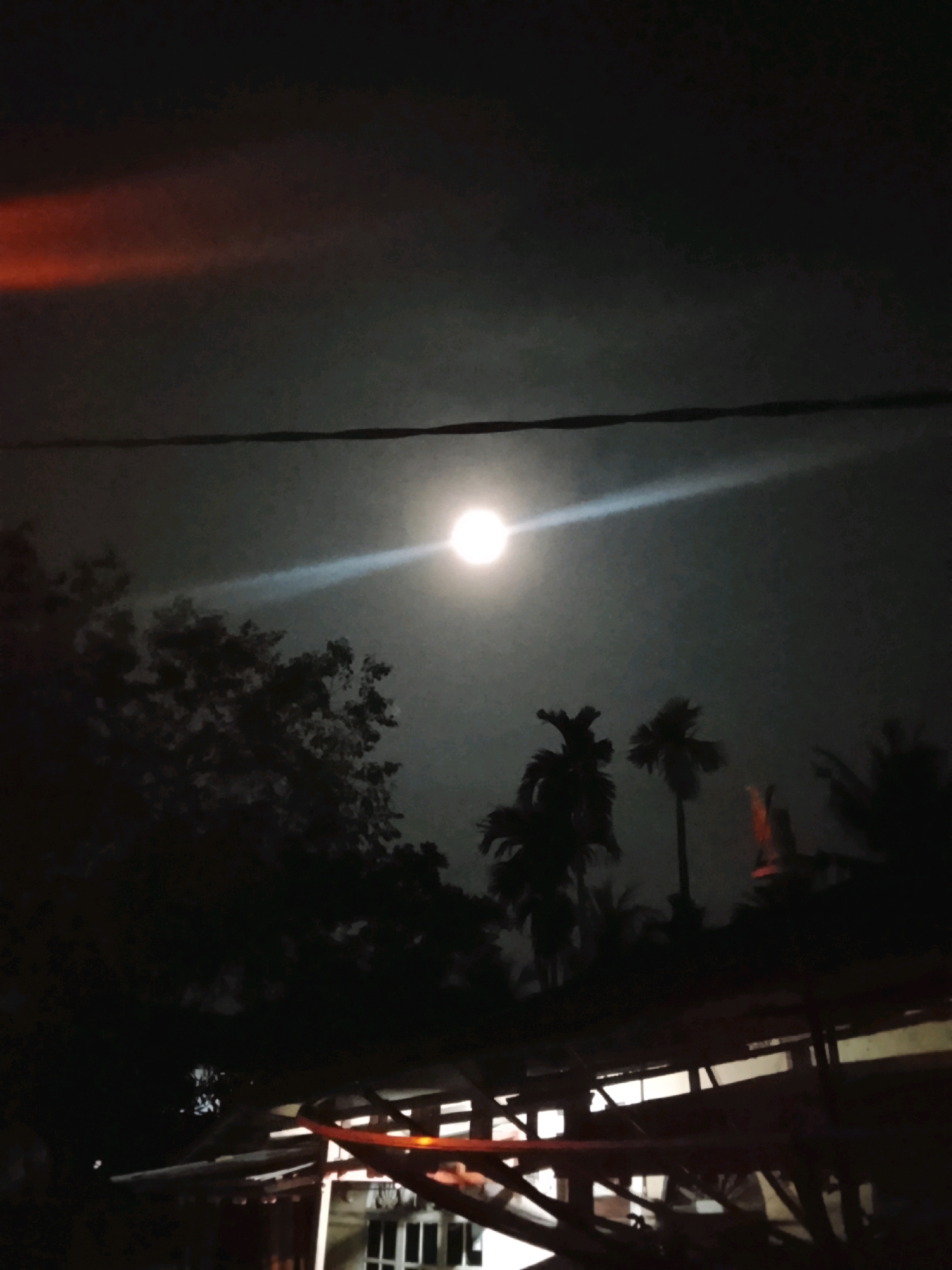 A nighttime photograph captures a bright moon illuminating a dark sky, with a faint orange glow visible in the upper left quadrant. Silhouettes of trees, including palm trees, are discernible against the sky. A power line stretches horizontally across the mid-section of the frame. In the foreground, a dark structure, possibly a building or rooftop with visible beams and supports, is present. The image appears to be taken in Lhokseumawe, Indonesia, given the presence of tropical vegetation like palm trees and the general ambiance consistent with a night scene in a coastal Indonesian city.