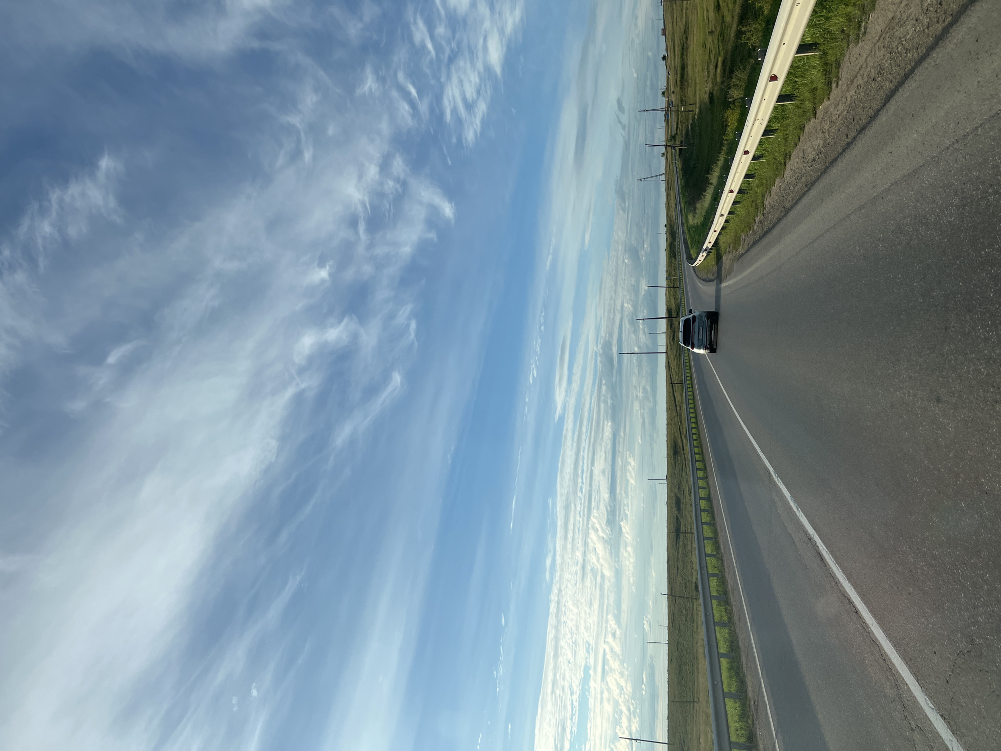 The image depicts a paved highway curving through a grassy, rural landscape under a bright blue sky with wispy white clouds. A silver SUV is traveling away from the viewer on the highway. A metal guardrail runs along the side of the road, separating it from the grassy fields. Utility poles are visible intermittently along the roadside. The scene appears to be an outdoor, daytime shot taken from a moving vehicle. The location is identified as Bolshoy Oyosh, Russia.
