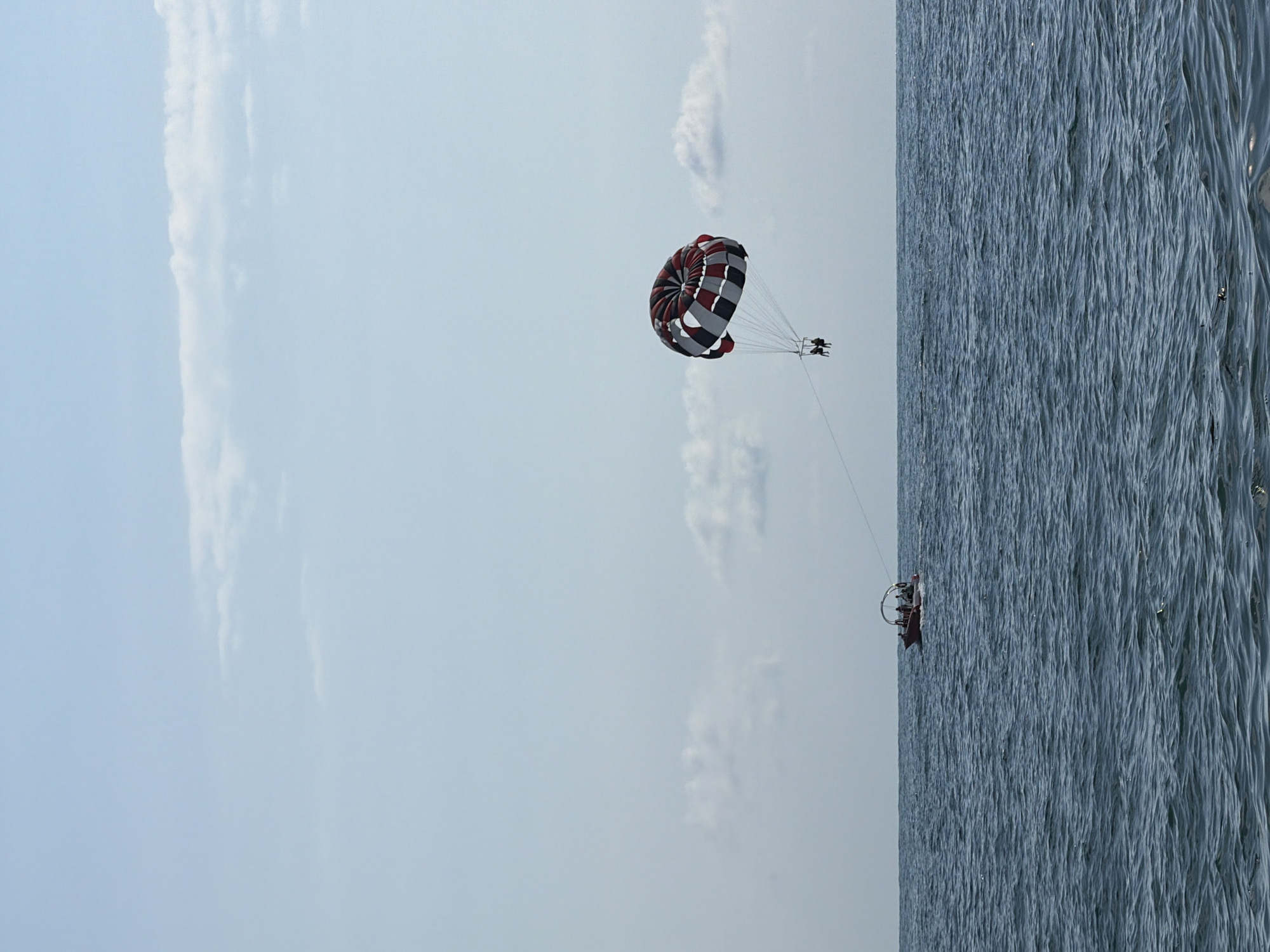 In the city of Sirius, Russia, a bright red, white, and black parasail is aloft in a clear sky dotted with a few wispy clouds. Below the parasail, two individuals are seated, suspended by the tow rope. Further in the distance, a small red boat navigates the deep blue, rippling waters of the ocean. The scene depicts a recreational activity taking place on a calm day with good visibility.