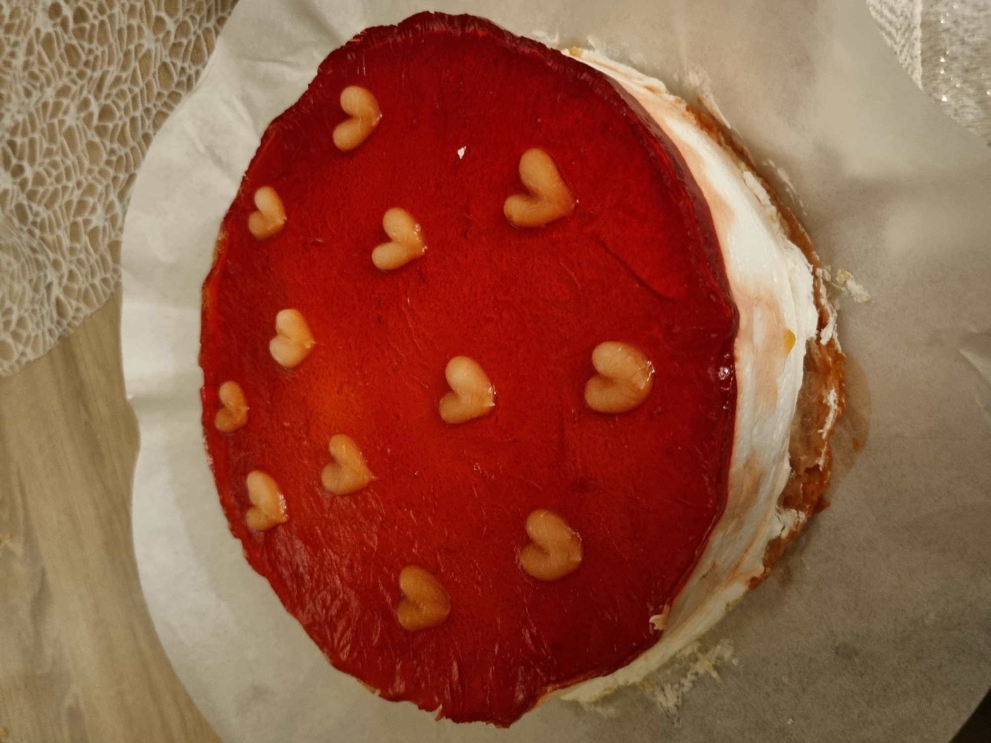 A top-down shot shows a cake with a bright red jelly topping, decorated with small, pale hearts arranged somewhat randomly. The cake is round and appears to have a layer of white frosting around the sides. It sits on a piece of parchment paper, which in turn rests on a wooden surface. In the upper left corner of the image is a lacy, textured fabric. The photograph does not provide information to indicate the location in Saarbrücken, Germany.