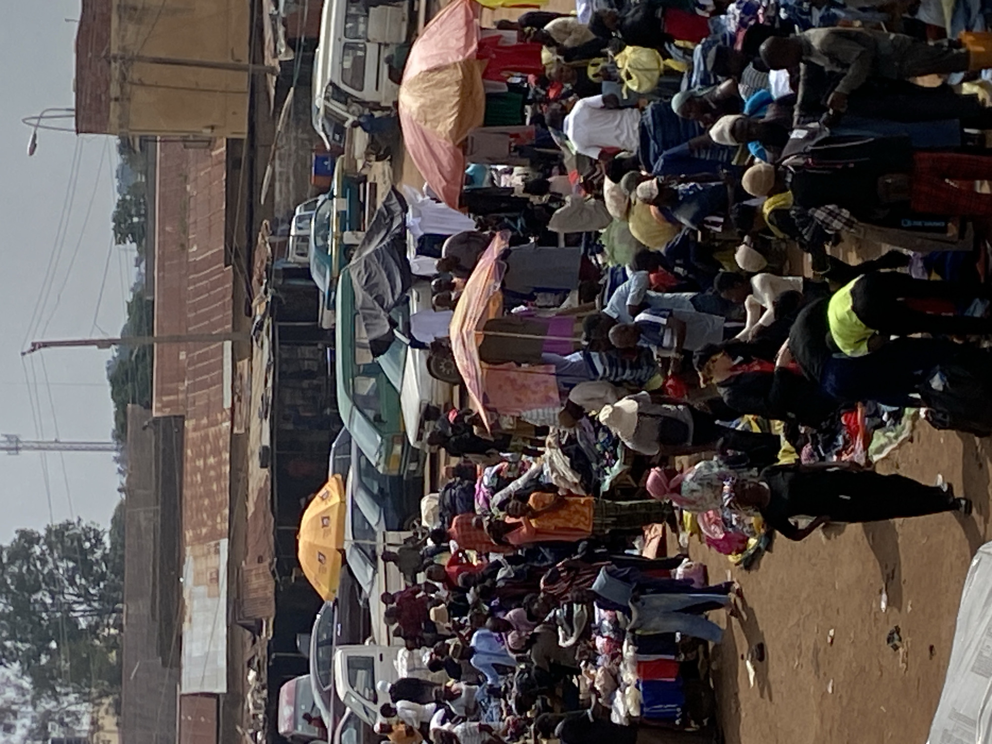 This is a vertical, medium-shot, outdoor photograph of a bustling marketplace in Bukuru, Nigeria. The scene is crowded with numerous people, likely vendors and shoppers, interacting amidst piles of goods, primarily clothing, displayed on the ground and on tables. Several market stalls and makeshift shelters with umbrellas and tarps are visible, providing shade from the overcast sky. Various vehicles, including cars and vans, are interspersed throughout the marketplace, suggesting it is a busy commercial hub. The background shows a mix of buildings with corrugated metal roofs, some appearing weathered, and trees, indicating an urban or semi-urban environment. The ground is dusty and uneven. The overall impression is one of lively commerce and activity.