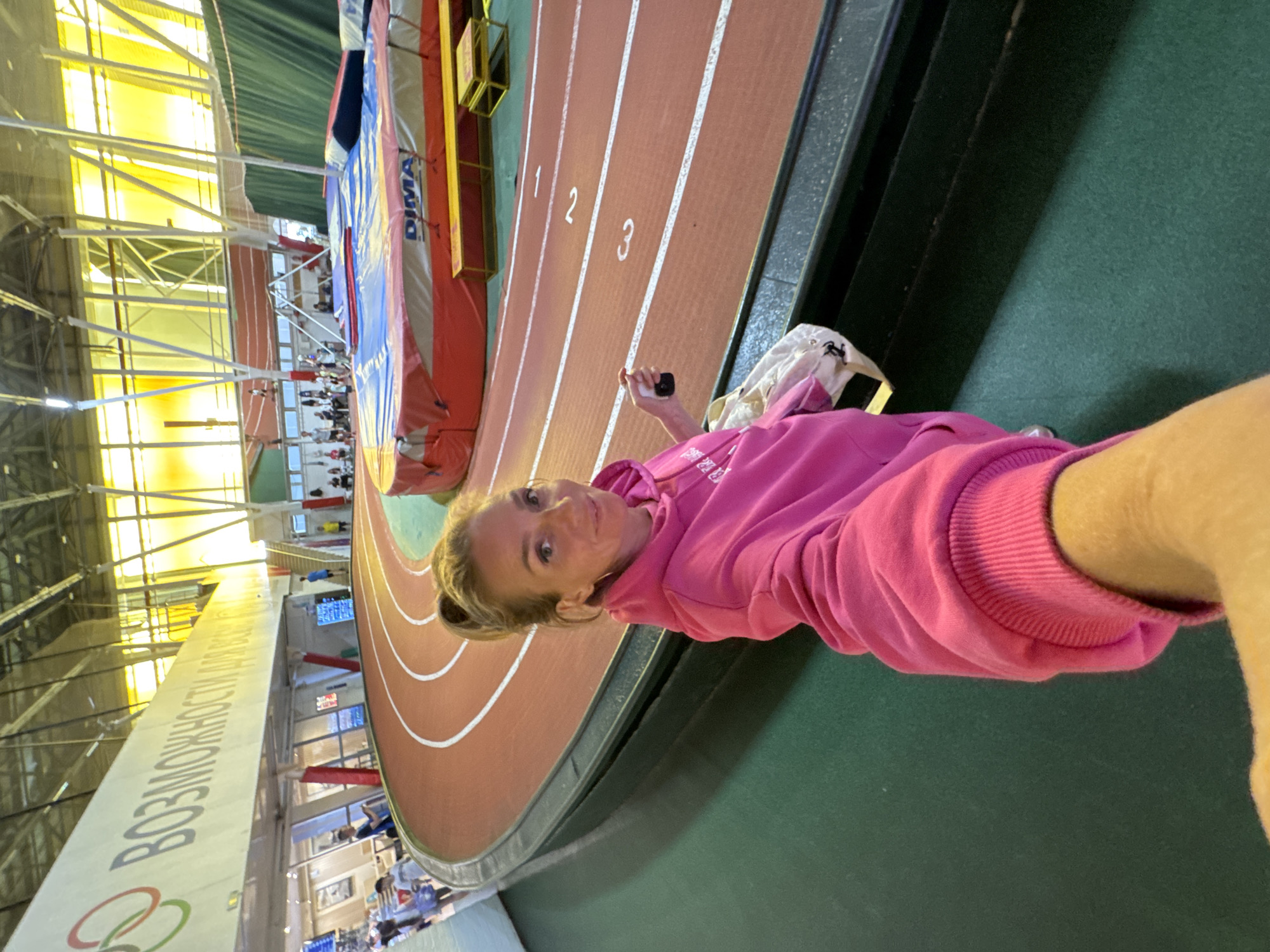 An indoor athletic stadium in Saint Petersburg, Russia, is depicted from a low-angle selfie perspective. A smiling woman with blonde hair tied in a ponytail, wearing a bright pink hooded sweatshirt, occupies the foreground, extending her right arm to capture the image. Her left hand holds a small black object, possibly a vape pen, and a white bag hangs from her left wrist. Behind her, a curved, three-lane running track with red surfacing and white lane markings numbered '1', '2', and '3' dominates the midground. Adjacent to the track is a dark green floor. In the background, a large crash mat branded "DIMA," typically used for high jump or pole vault, is visible with yellow benches nearby. An elevated walkway or mezzanine stretches above the track, featuring a long white sign with the Russian word "ВОЗМОЖНОСТИ" (Opportunities) and a red, green, and blue three-ring logo, reminiscent of a sports organization emblem. Numerous individuals are scattered throughout the background, on the track level and the upper walkway. The facility is illuminated by natural light through yellow-tinted windows and overhead artificial lighting, with exposed metal trusses supporting the ceiling.