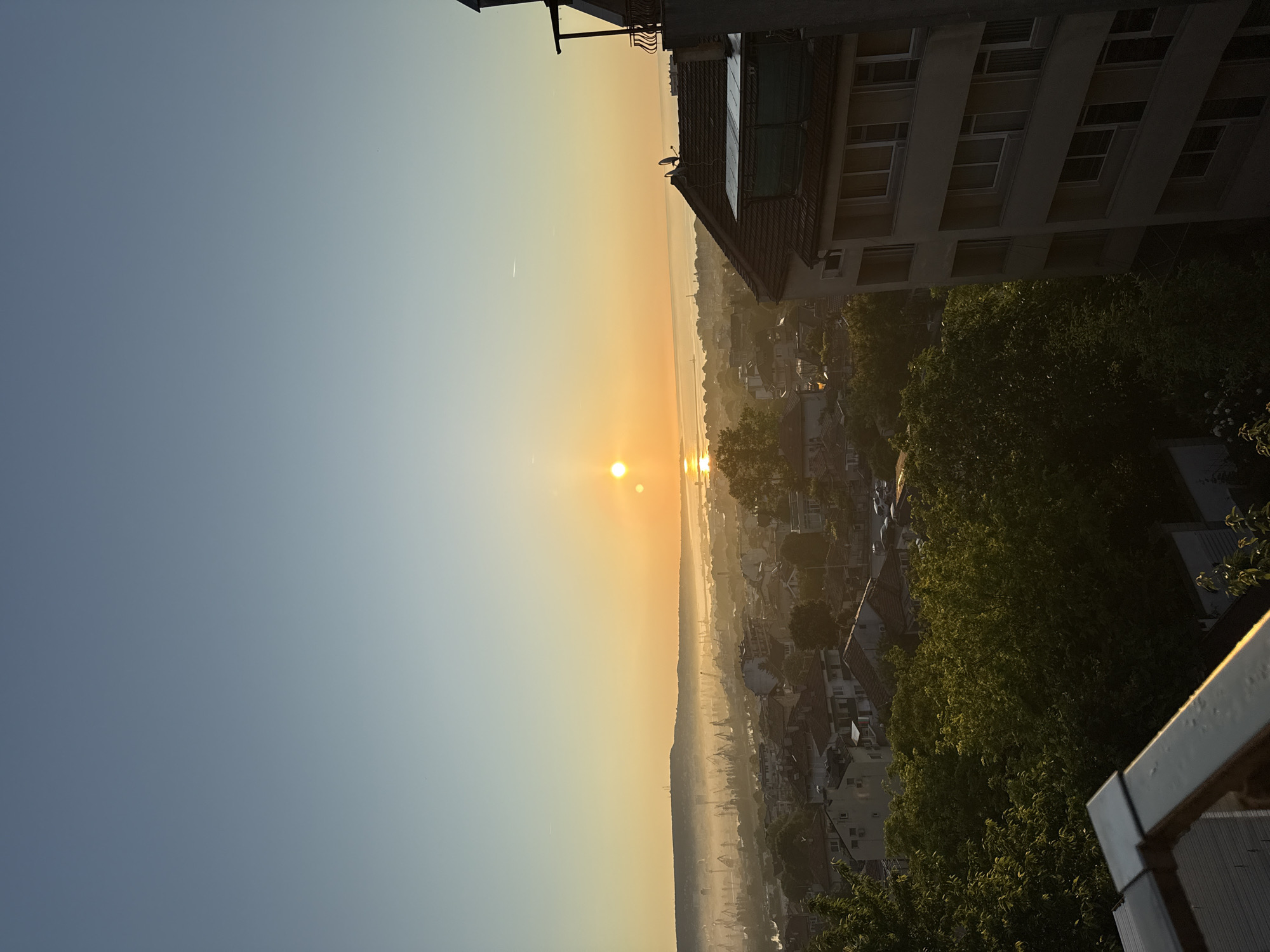 This is a high-angle, full-shot photograph of a cityscape at sunrise. The sky is a gradient of pale blue to soft yellow as the sun rises over a hazy horizon. The sun itself appears as two distinct, bright orbs. In the foreground, a portion of a balcony railing is visible, along with lush green trees. Beyond the trees, a dense urban area with red-tiled roofs and residential buildings unfolds. In the midground, a port or industrial area is discernible, characterized by numerous cranes and structures, partially obscured by morning mist. The scene is one of a waking city bathed in the warm glow of early morning light.