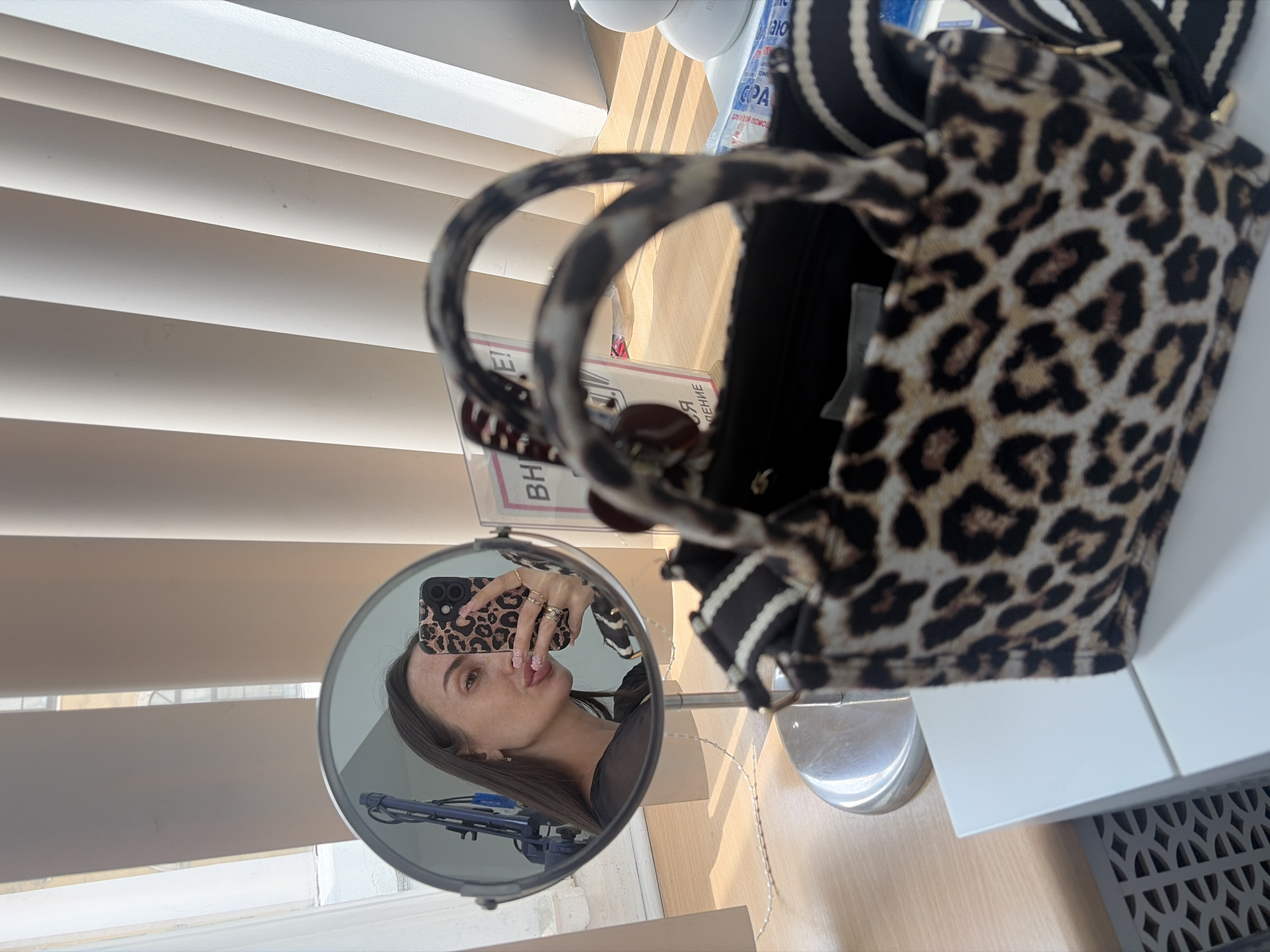 A woman, reflected in a circular mirror, takes a selfie with her phone. She is wearing a black top and has long, dark hair. Her makeup includes lipstick and eyeliner. A leopard-print handbag sits in the foreground. The background appears to be an indoor setting with blinds and a wooden surface. The image does not contain any specific landmarks or clues to indicate a location within Saint Petersburg, Russia.