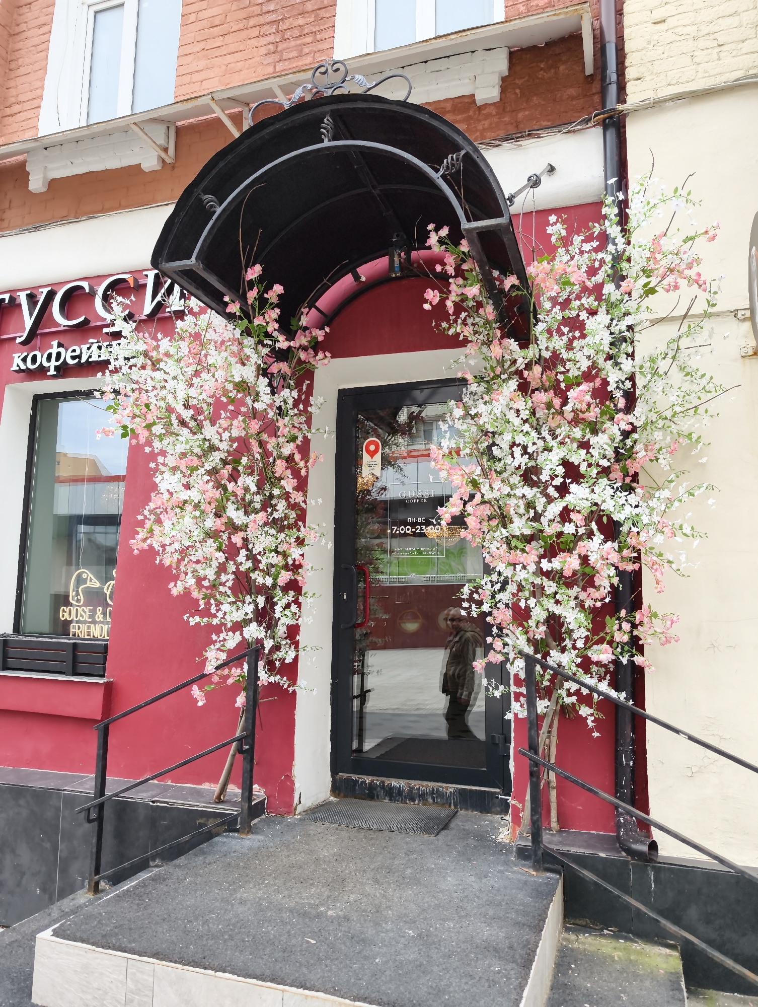 Here is a detailed description of the image:
**Scene:** The image shows the entrance to "GUSSI COFFEE," a coffee shop in Saratov, Russia.
**Location:** The entrance is decorated with two large floral arrangements featuring white and pink blossoms.
**Architecture:** The building has a red facade on the left and a light beige facade on the right of the entrance. The doorway is framed in red, with a black awning over the entrance. Black metal railings flank the steps leading to the door.
**People:** The reflection of a person is visible in the glass door.
**Objects:**
* **Signage:** A sign on the window reads "Русси кофейн" (Russian Coffee) and a smaller sign under it says "GOOSE & CO FRIENDLY". A decal shows a pair of geese. Opening times are displayed on the door as being 7:00-23:00.
* **Exterior Features:** A window is visible to the left of the door.
* **Material**: The entry has tiled steps and a dark mat.
**Environment:** The scene is outdoors during daylight. The building appears to be in a bustling area, given the presence of other buildings nearby.
**Overall Impression:** The coffee shop appears inviting and well-maintained. The floral decorations provide a welcoming aesthetic.