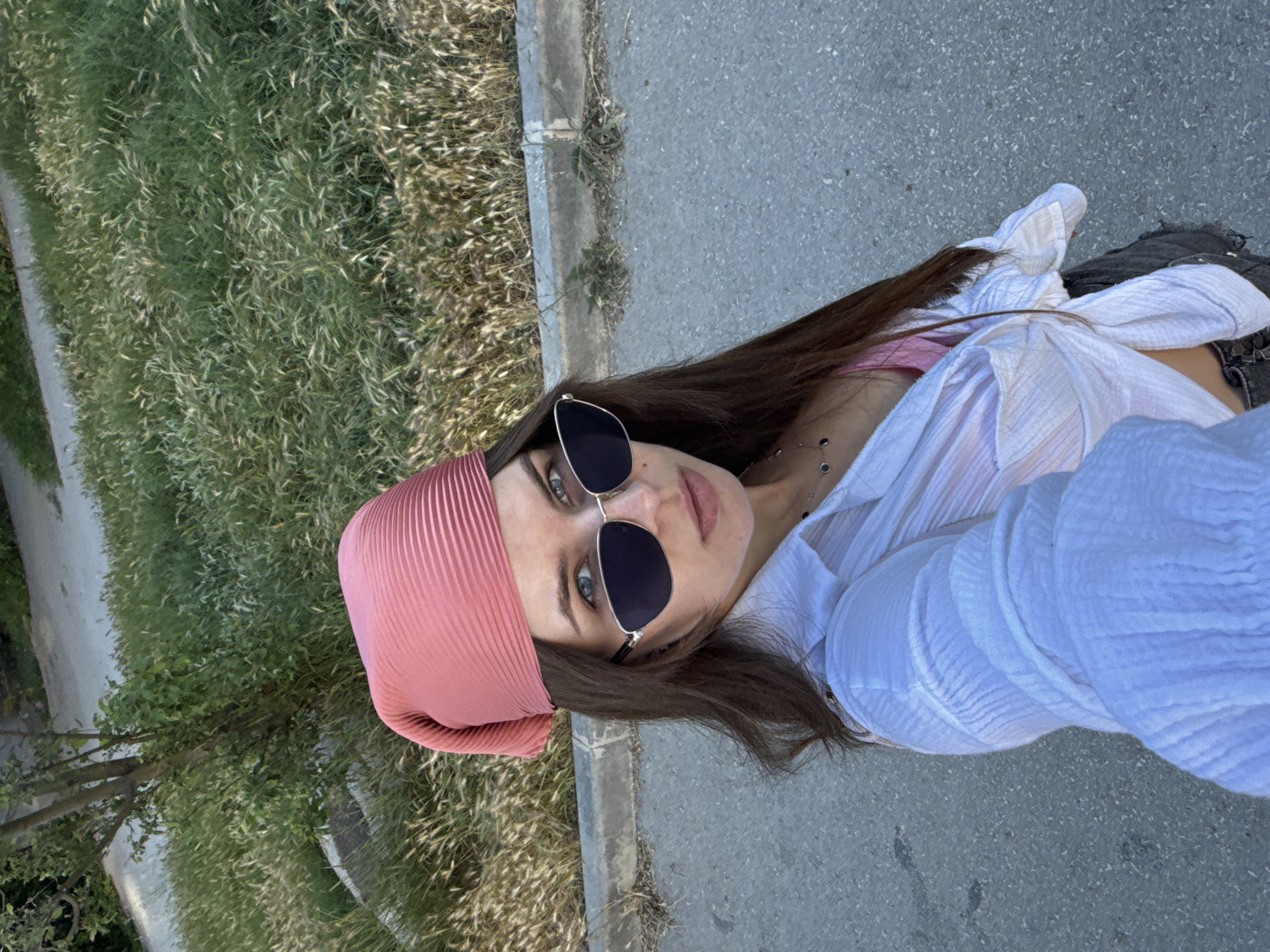 A woman is taking a selfie outdoors, standing on a paved surface. She is wearing a pink headscarf, sunglasses, and a white unbuttoned shirt over a pink top. Her dark hair is long and falls around her shoulders. She is wearing a necklace with a dark pendant. In the background, there is a grassy area with some trees and a concrete curb. The image is taken from a high angle.