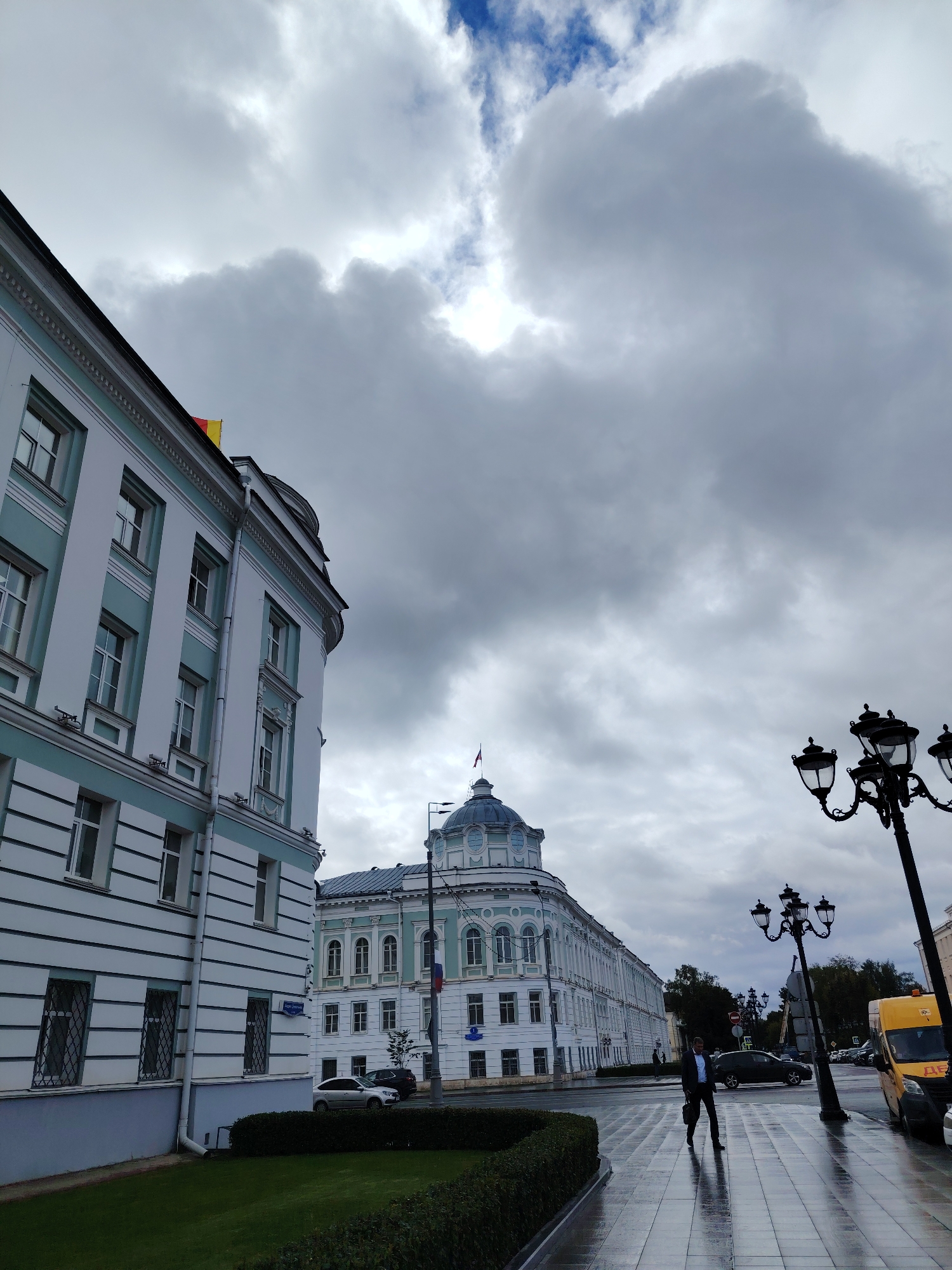 The image shows a street scene in Tver, Russia, with the Tver Imperial Palace visible in the background. The sky is overcast with dramatic clouds, and the wet pavement suggests recent rain.
In the foreground, a light blue and white building with multiple windows lines the left side of the street. A neatly trimmed hedge separates the sidewalk from a grassy area.
On the right side of the image, a man in a dark suit and briefcase walks across the wet sidewalk. A vintage-style street lamp with three globes stands tall, its silhouette stark against the sky. Further down the street, another street lamp and a portion of a yellow vehicle are visible.
The Tver Imperial Palace, a grand building with a dome and arched windows, is situated in the middle ground. A flag flies from its roof. The street itself appears to be a main thoroughfare with parked cars and a bus. The architecture is characteristic of historic European cities.
