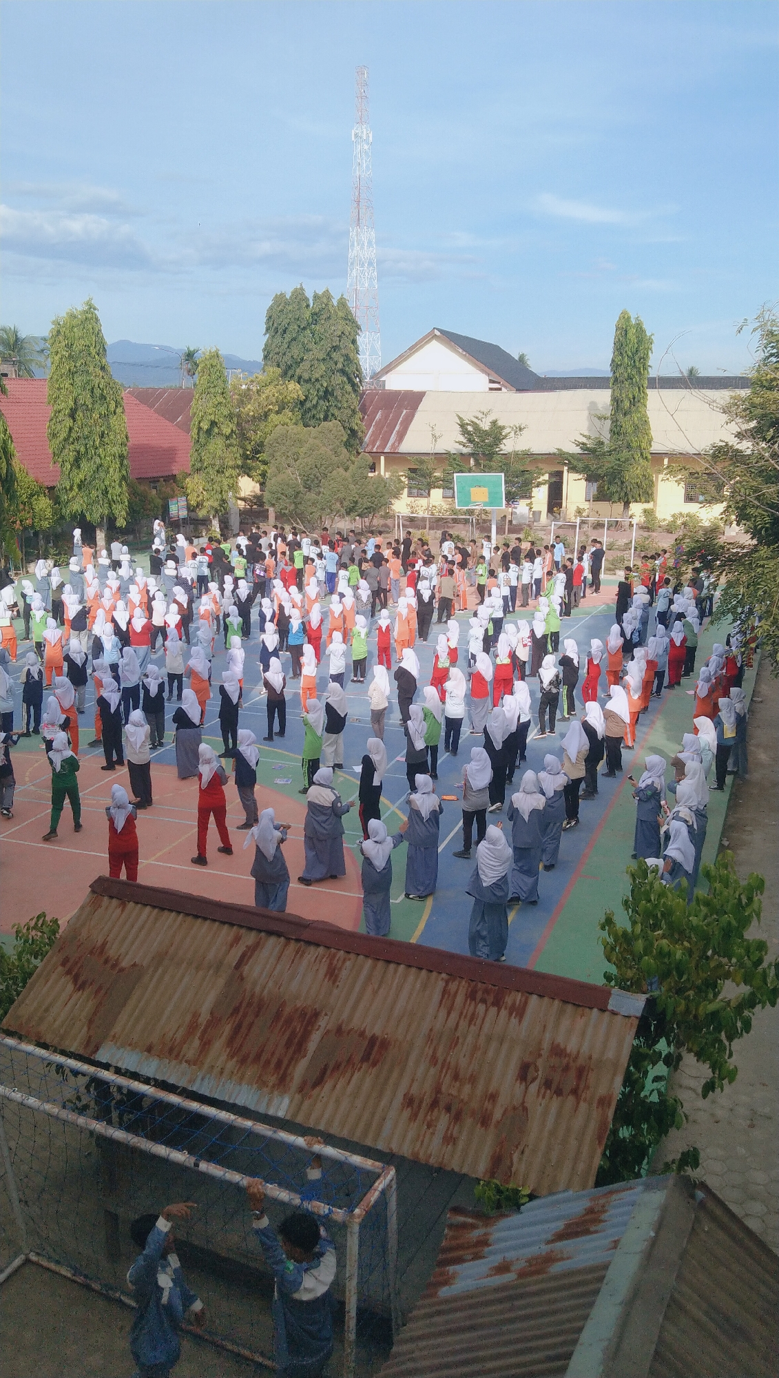 This image captures a wide-angle view of an outdoor morning exercise or assembly at a school in Rhieng Blang, Indonesia. A large group of students, predominantly female and wearing hijabs, are positioned on a sports court with marked lines for various games, possibly volleyball or basketball, indicated by a hoop visible in the background. The students are arranged in multiple rows, facing a common direction, and appear to be performing synchronized movements, suggesting an organized activity. Their attire varies, with many wearing colorful uniforms, including orange, white, blue, and gray.
The environment surrounding the court includes school buildings with light-colored walls and brown-tiled roofs. Tall, slender evergreen trees are scattered around the school grounds, and in the distance, a prominent telecommunications tower stands against a pale blue sky with scattered clouds. The foreground of the image is dominated by the roof of a low-lying structure, possibly a shed or covered area, constructed with corrugated metal that shows signs of rust. Two young individuals in blue uniforms are visible in the lower left corner, interacting with a goalpost net, potentially preparing for or observing the activity. The overall scene depicts a typical school day scene in a tropical or subtropical climate, with the natural light suggesting it is either early morning or late afternoon.