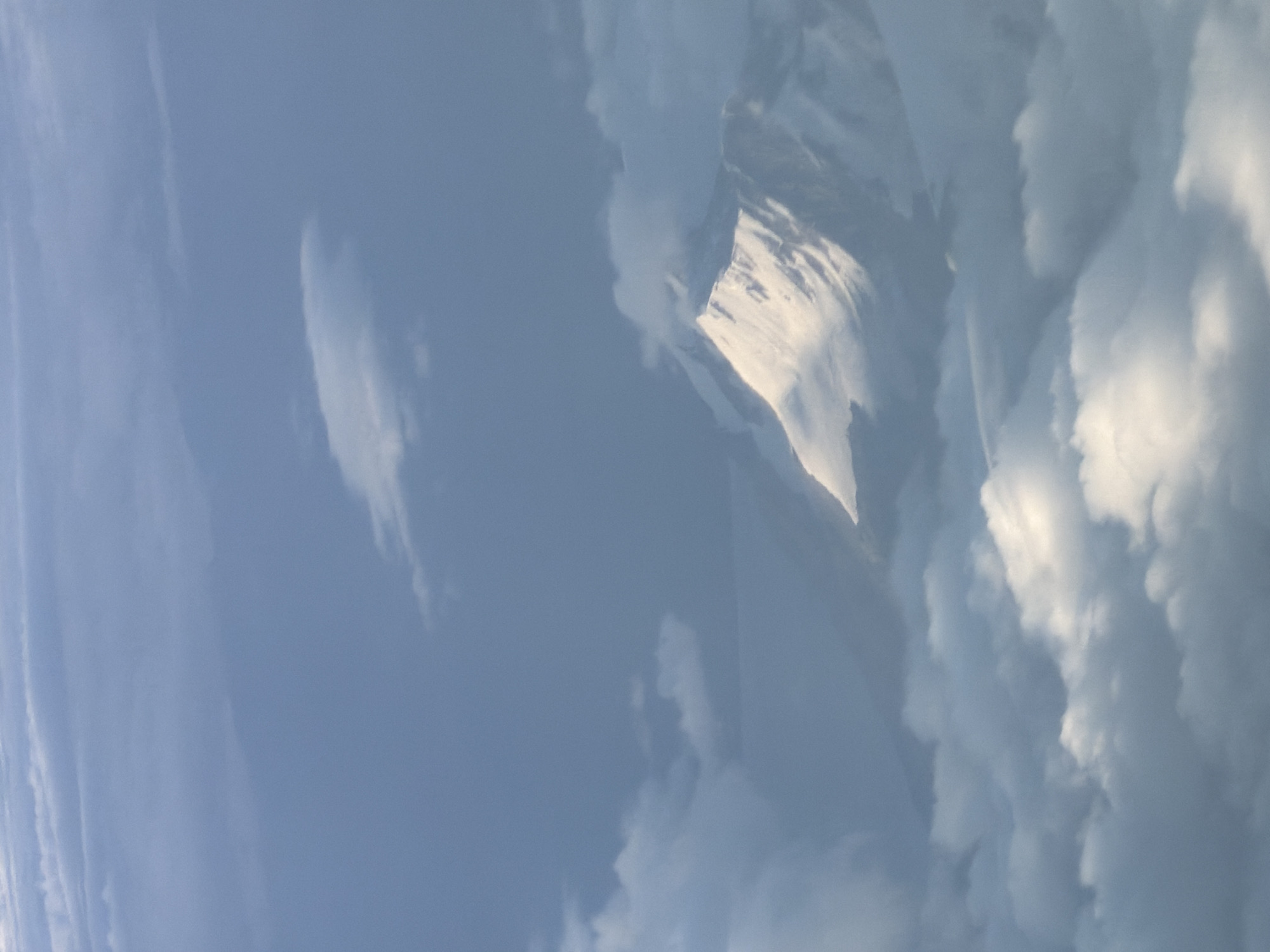 An aerial view captures a snow-capped mountain peak emerging from a blanket of clouds. The mountain, illuminated by sunlight, displays a sharp, angular profile with visible ridges and slopes. Surrounding the mountain are numerous white, fluffy clouds, some of which partially obscure the lower portions of the peak. The sky above is a soft blue, with some wispy cloud formations. The image appears to be taken from a high altitude, possibly from an aircraft, looking down on the mountain range. The location in Hiranagar, India, is not identifiable from the visual information alone, as the image depicts a natural landscape.