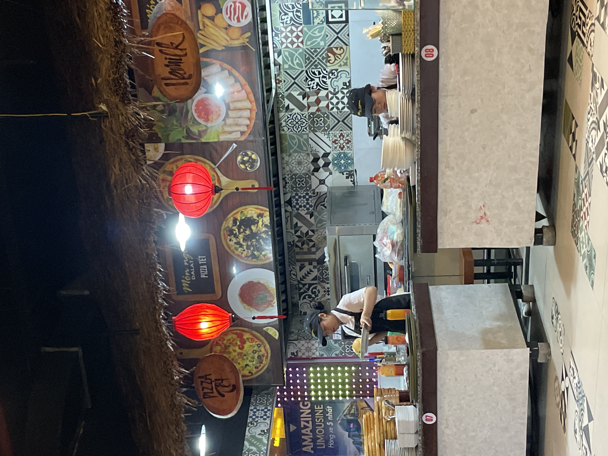 This image depicts an indoor food service area, likely a restaurant or food court, situated in Da Lat, Vietnam. The ceiling features a decorative thatched straw covering, from which two glowing red spherical lanterns are suspended. Two female individuals, both wearing black caps, white shirts, and aprons, are positioned behind separate service counters. The person on the left, identified by counter number "07," holds a plate bearing a burger-like item. The person on the right, at counter "08," is leaning forward, partially obscured by stacks of white dishes, engaging in preparation or arrangement. Both counters are light-colored with dark brown trim. Bottles of condiments are visible on the left counter. The back wall above the counters is tiled with a diverse array of geometric and abstract patterns. Above the tiling, large graphic murals display various food items, including pizzas, spring rolls with dipping sauce, and french fries. Several signs are present: a wooden oval sign inscribed "PIZZA" and additional text, a blackboard-style sign reading "Món ngon DALAT / PIZZA TẾT," and another wooden oval sign with the word "Hoanilk." On the far left, a vertically oriented blue sign with yellow lights advertises "AMAZING LIMOUSINE / Hàng xe 5 nhất." The floor is adorned with patterned tiles featuring intricate geometric and floral designs.