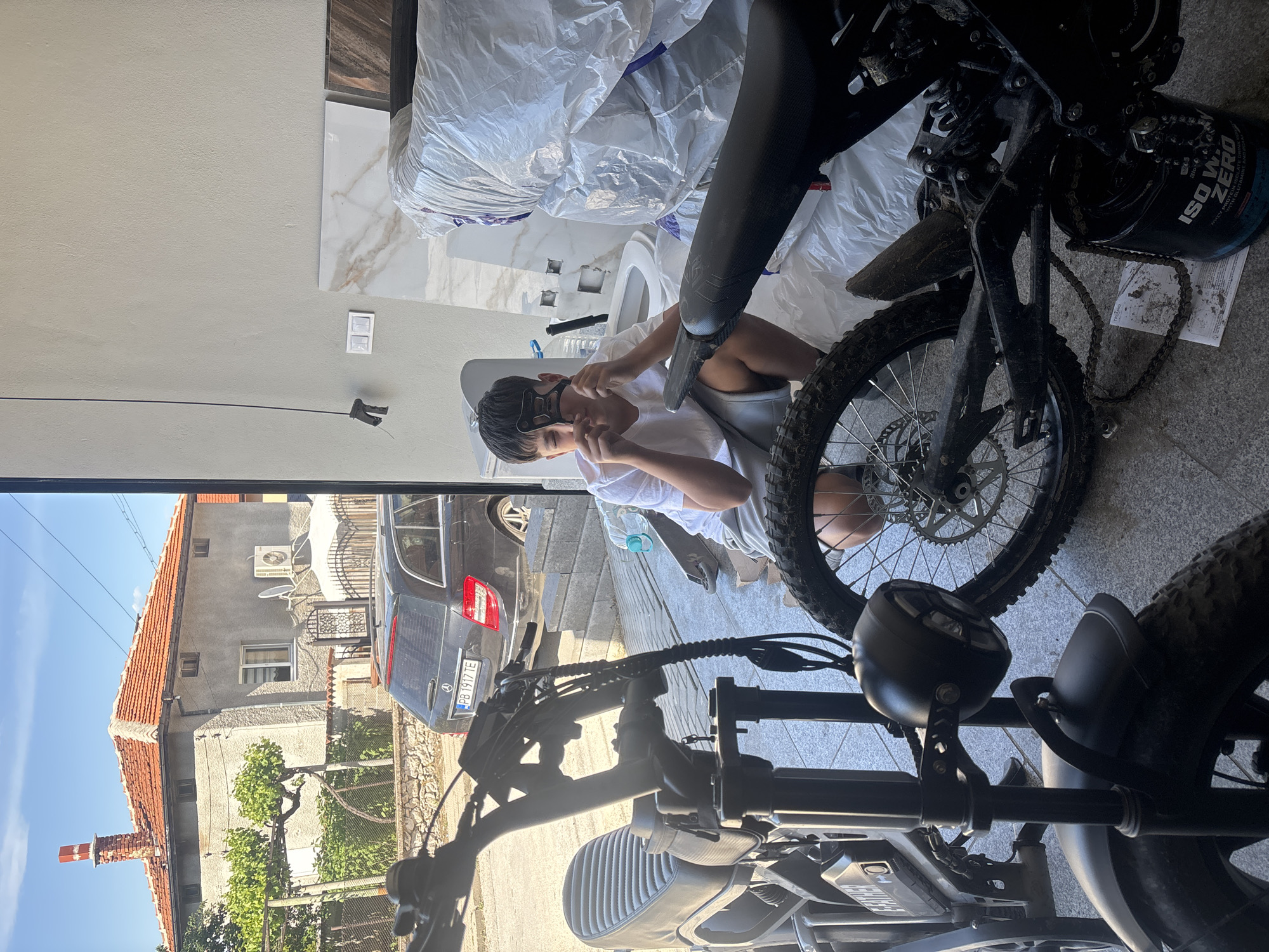 The scene is a partially obscured view through a doorway or window, showing both an interior and exterior space. Inside, a young person wearing a white t-shirt and grey shorts is seated near what appears to be a toilet bowl with a construction site of a new bathroom. They are holding and adjusting a black object near their face. Around them are construction materials, including large bags of white material. There is a chain for a dirt bike on the floor.
In the foreground, parts of two black dirt bikes are visible.
The exterior view includes a car parked in front of a residential building with a red tiled roof. The car has a visible license plate that reads "PB 1917 TE". The image suggests an outdoor area with trees and a blue sky.
The location, based on the license plate code "PB", is likely in the area of Pazardzhik in Bulgaria. The image appears to be taken in or around the city of Asenovgrad, Pazardzhik Bulgaria, as it's where the photo was taken.