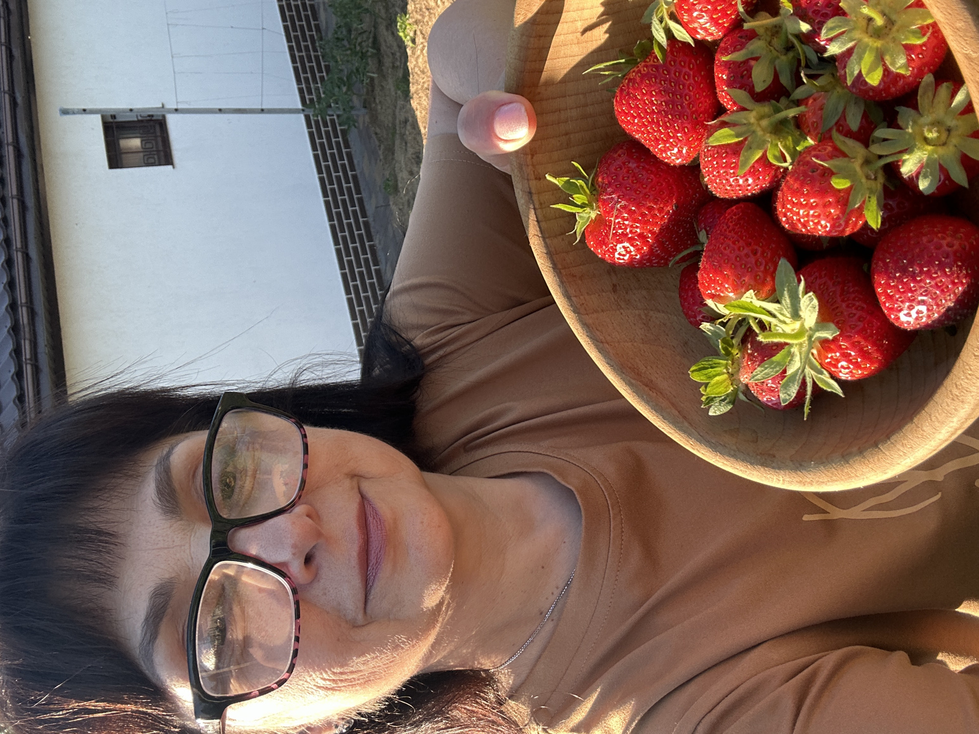 In Hnarovske, Ukraine, a woman with dark hair and glasses, wearing a tan t-shirt, holds a wooden bowl filled with freshly picked strawberries. She is positioned in the foreground, with a white building and a brick wall visible in the background, suggesting an outdoor setting, possibly a garden or yard. The sunlight casts a warm glow on the scene, highlighting the vibrant red of the strawberries and the woman's features.