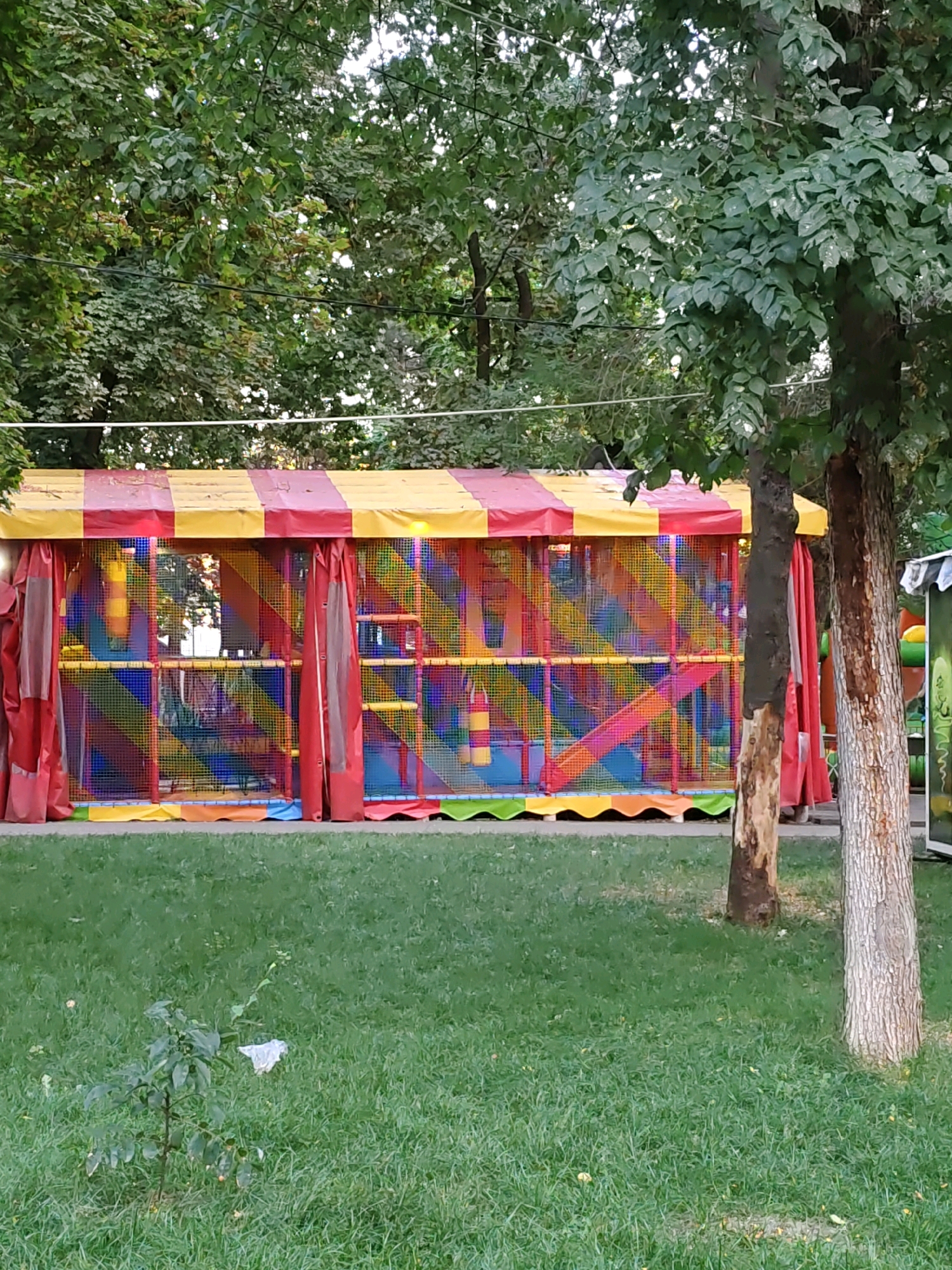 A brightly colored inflatable playground structure, covered by a striped red and yellow tent, is visible in a grassy outdoor area. The playground features netting and various brightly colored slides and climbing structures. Red fabric drapes hang from the sides of the tent. The playground is surrounded by green grass, with several trees in the foreground and background. The overall scene suggests a public park or recreational area. The location is a playground in Rostov-on-Don, Russia.