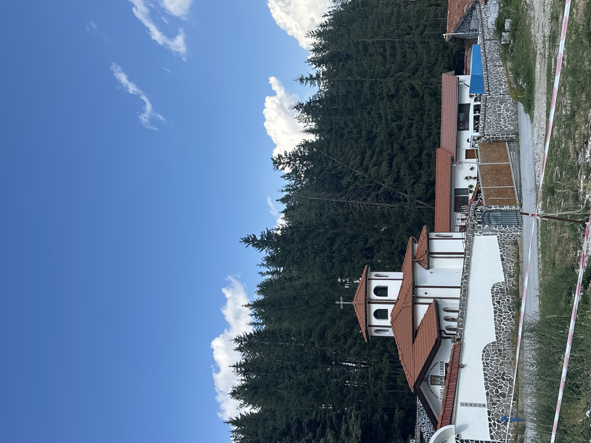 The image depicts a white building with red-tiled roofs, possibly a church or monastery, set against a backdrop of a dense evergreen forest under a bright blue sky with scattered white clouds. The building features arched windows and a cross on its highest point. A stone and white wall surrounds the property, with an entrance gate visible. The ground in front of the wall is covered with gravel and dry grass, with pink and white caution tape running horizontally across the lower part of the frame. The overall scene suggests a rural or semi-rural setting, potentially in the vicinity of Smolyan, Bulgaria, given the architectural style and the prevalence of conifer forests in the region.