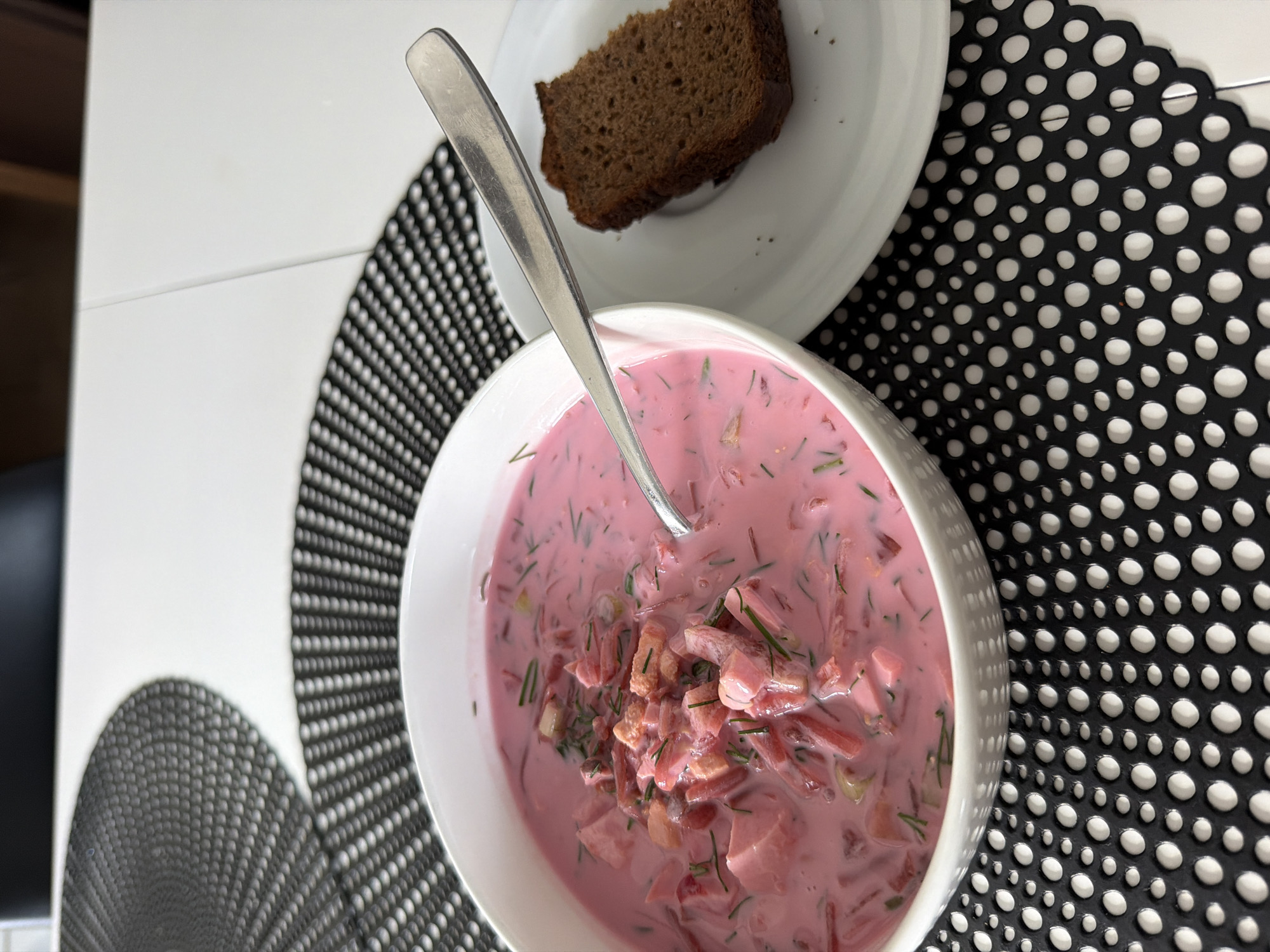A high-angle shot captures a bowl of pink cold beet soup, commonly known as 'šaltibarščiai' or 'chłodnik', with a spoon resting within it. The soup, vibrant in color, contains finely chopped ingredients such as beets, possibly cucumber, and fresh green dill. Beside the soup, on a small white plate, sits a thick slice of dark, dense bread, likely rye. The meal is presented on a white tabletop, partially covered by two decorative placemats. The main placemat, positioned under the soup and bread, is black with an intricate pattern of various-sized white circular holes or dots. A second, similar placemat is partially visible in the bottom left corner. The setting suggests a casual meal in a home environment, consistent with the provided location context of Baloži, Latvia, where such a soup is a popular dish. No people or visible text are present in the image. The lighting appears natural, indicating daytime.
