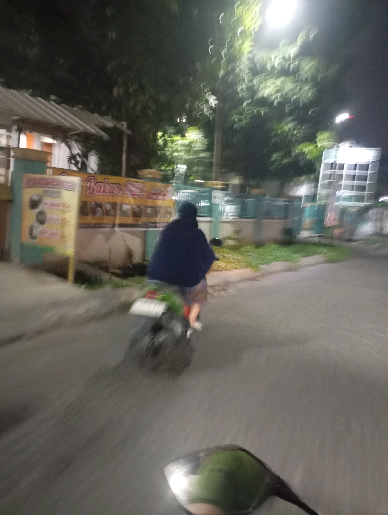 A night time, blurry, medium-shot photograph captures a person riding a motorcycle through a street in Babelan, Indonesia. The rider, wearing a dark blue hijab and a patterned skirt, is positioned in the center of the frame, moving away from the camera. The motorcycle, a dark color with a green fender, is also visible. The background features a roadside stall with a yellow banner displaying text and images, likely indicating food items for sale. Adjacent to the stall is a white structure with a corrugated roof. Further back, trees with illuminated foliage line the street, suggesting the presence of streetlights. On the right side of the image, a tall white structure with a grid-like pattern is visible. The road surface appears wet, reflecting the ambient light. The overall impression is of a transient, urban scene at night.