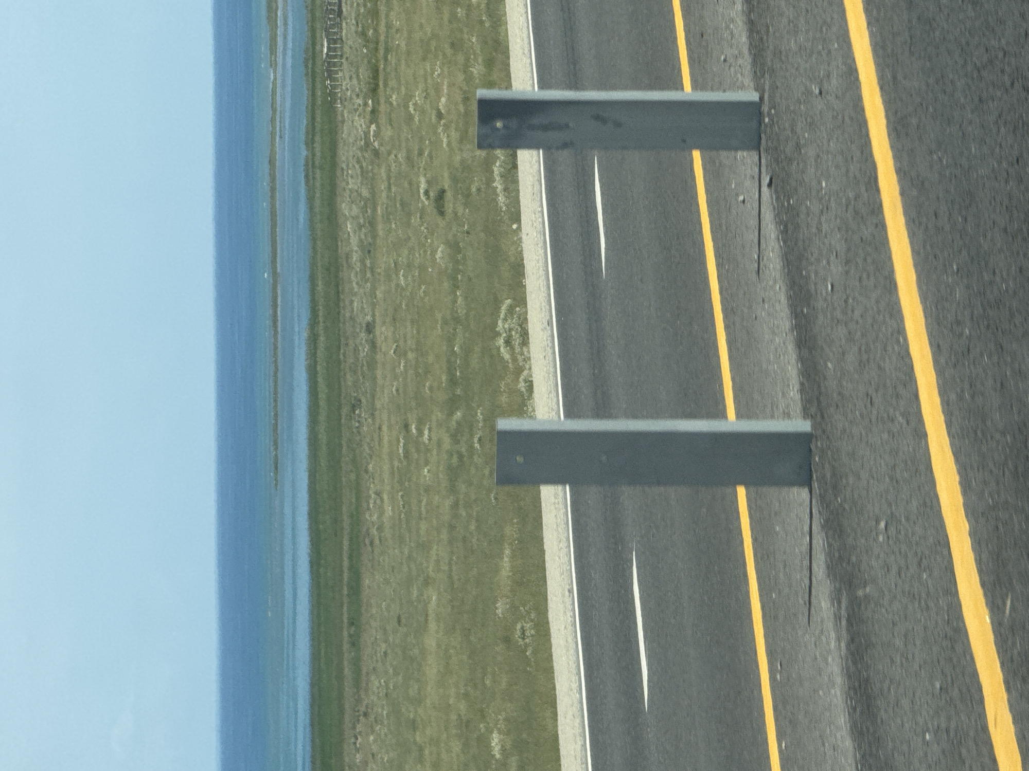 The image displays a roadside scene with a body of water and grassland in the background. In the foreground, a paved road with yellow and white lane markings is visible. Two gray metal posts are positioned on the right side of the road, casting shadows. The landscape beyond the road consists of dry, green grassland, with a body of water and a light blue sky in the distance. The overall impression is that of a natural, open environment, possibly in a rural or undeveloped area. There are no people or specific landmarks visible that would definitively place the scene in Kara-Talaa, Kyrgyzstan.