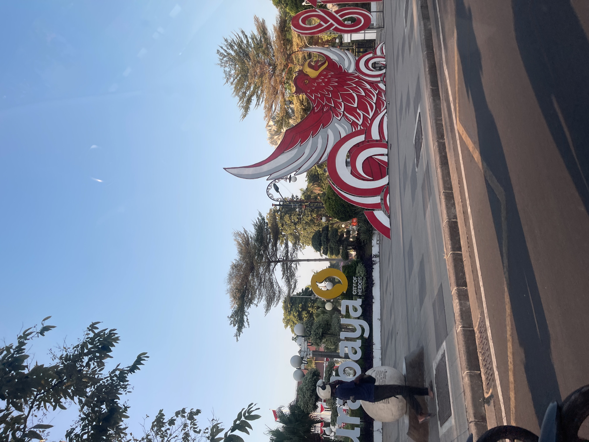 The image shows a street scene in Surabaya, Indonesia, with a large outdoor art installation. In the foreground, a person wearing a dark shirt and dark pants is walking past a large white spherical sculpture. Behind them, a prominent white sign spells out "SURABAYA," with "CITY OF HEROES" written below a yellow circular logo. To the right of the sign is a large, stylized red and white sculpture resembling a mythical bird or garuda, with its wings spread. Further to the right, large red numerals "8" and "7" are visible. The background features several trees, streetlights, and a clear blue sky. The street in the foreground has a curb and road markings, with shadows cast by unseen objects, suggesting it is daytime. The overall setting appears to be a public space or plaza in Surabaya.