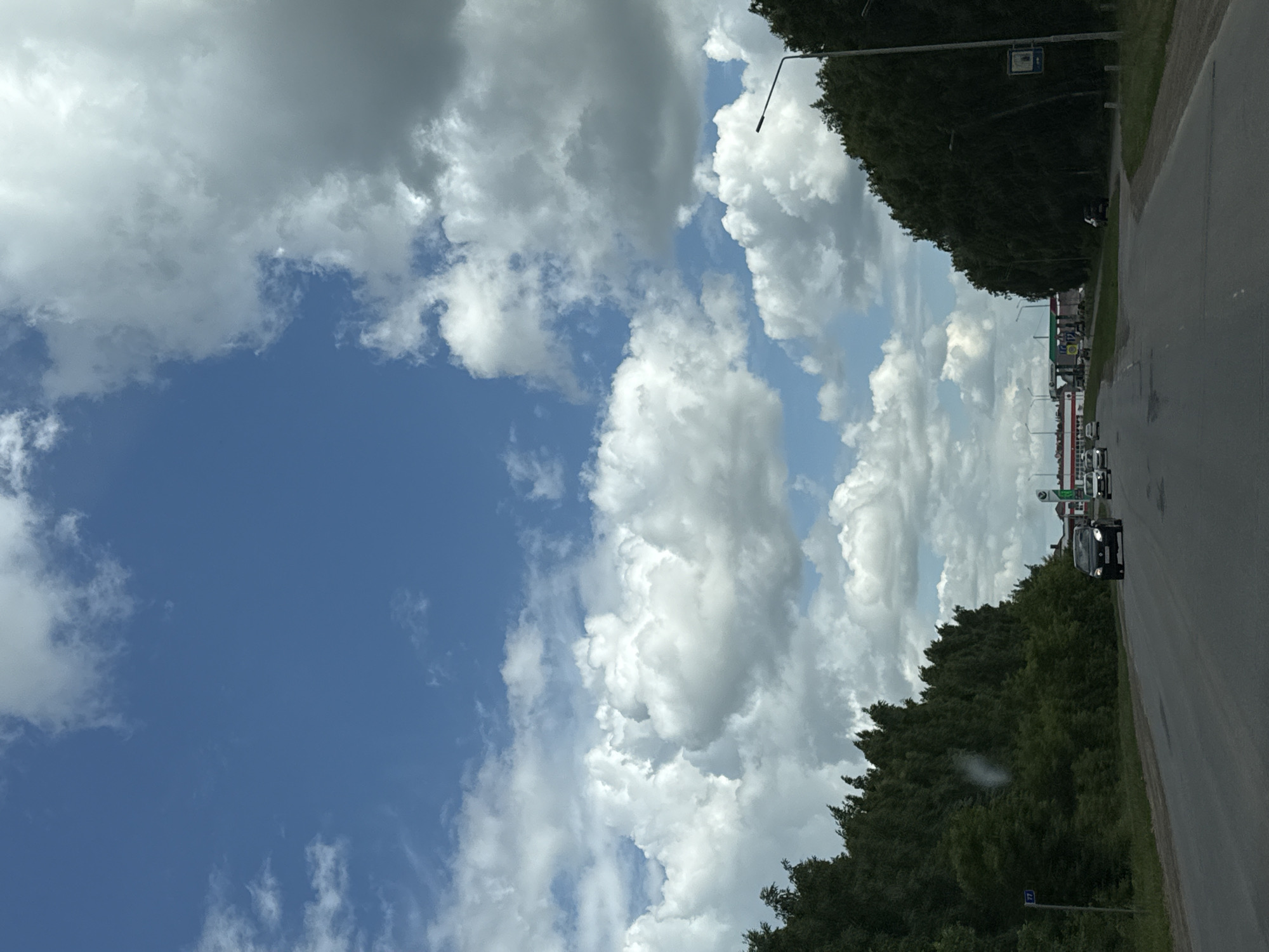 The image depicts a highway scene in Nurkeevo, Russia, under a partly cloudy sky. The road, paved with asphalt, curves slightly to the right. A line of dark green trees flanks both sides of the road, with the trees on the left appearing denser and taller. Several cars are visible on the highway, traveling in the same direction as the viewer. On the left side of the road, a blue street sign with the number "77" is present. In the distance, a commercial area with gas stations and buildings can be observed. A street light stands on the right side of the road, near the edge of the trees. The sky is a mix of bright blue and large, white and gray clouds.