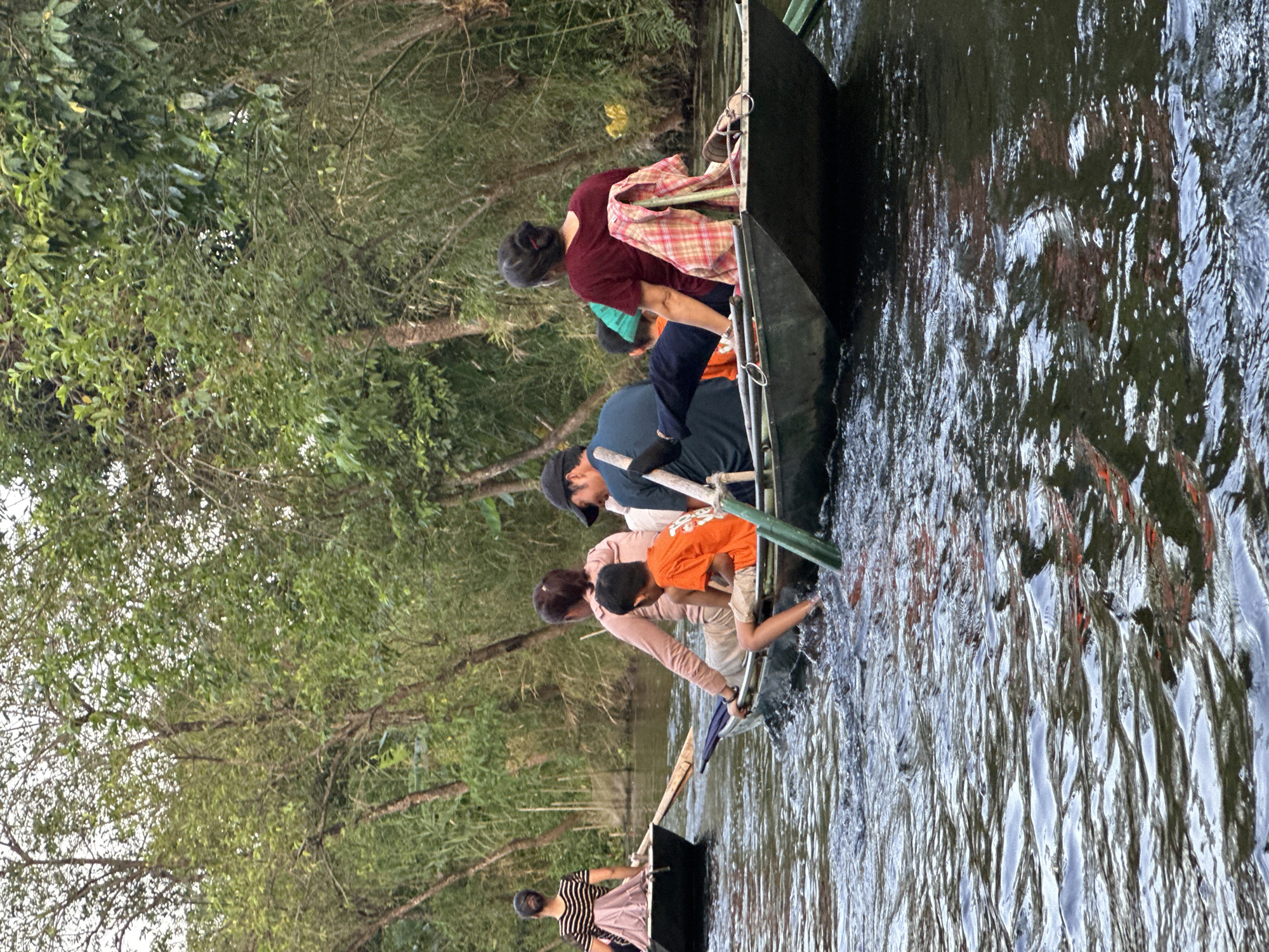 Two narrow, flat-bottomed boats are navigating a narrow, dark waterway lined with lush green trees and reeds, likely in the vicinity of Nam Hoa Lư Ward, Vietnam. In the foreground boat, four individuals are seated: a man wearing a dark shirt and a black cap is rowing with a wooden oar. Beside him, a woman in a maroon shirt has a plaid blanket draped over her shoulder. A young boy in an orange t-shirt is seated in the middle of the boat, and another person, partially obscured, is seated in front of him. In the boat further in the distance, a woman with her hair in a bun is visible from behind, wearing a striped t-shirt and a light pink skirt. She is also in a boat with another person in front of her. The water is rippling, reflecting the surrounding greenery and the boats.