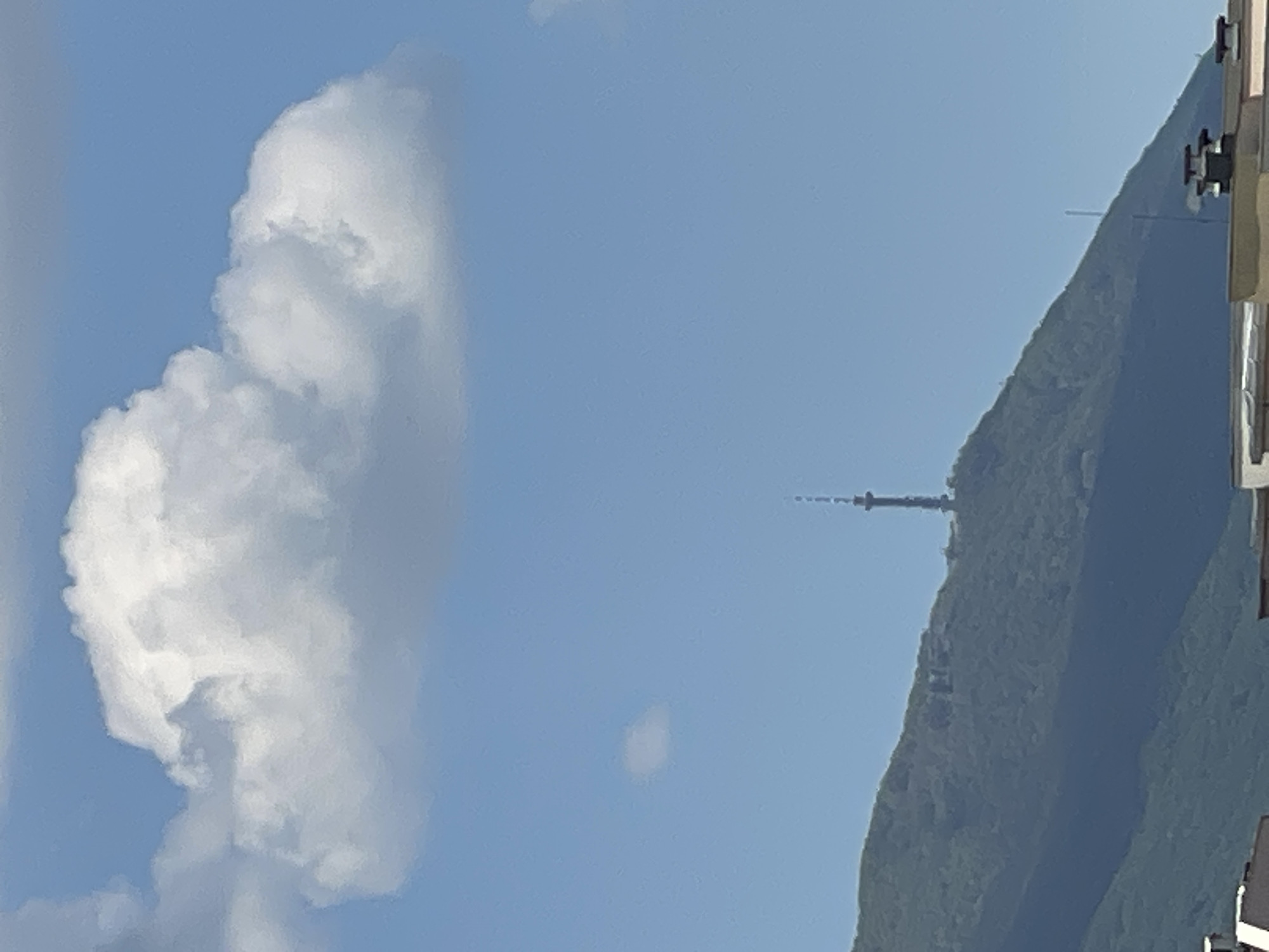 The image depicts a daytime scene with a large, fluffy white cloud in the left portion of the frame against a light blue sky. To the right, a forested hill or mountain slope is visible, with a tall transmission tower or communications antenna situated on the ridge. Some building parts appear on the right edge of the image. The location is likely Sofia, Bulgaria as the tower on the hill resembles Kopitoto TV Tower.