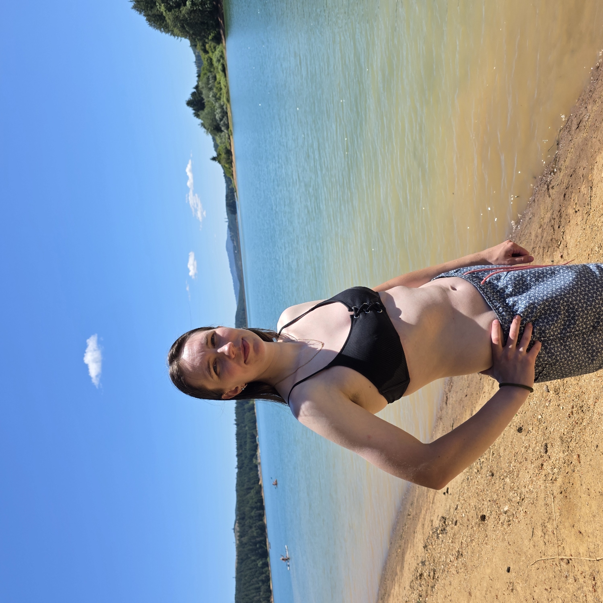 A woman stands on a sandy beach with a calm, blue lake behind her. She is wearing a black bikini top and patterned swim trunks. Her left hand is on her hip, and her right arm is bent. The background shows a tree-lined shore and hills in the distance under a clear blue sky with a few scattered clouds. There are some people on paddleboards in the lake. The location is the city of Cherry Grove, United States.