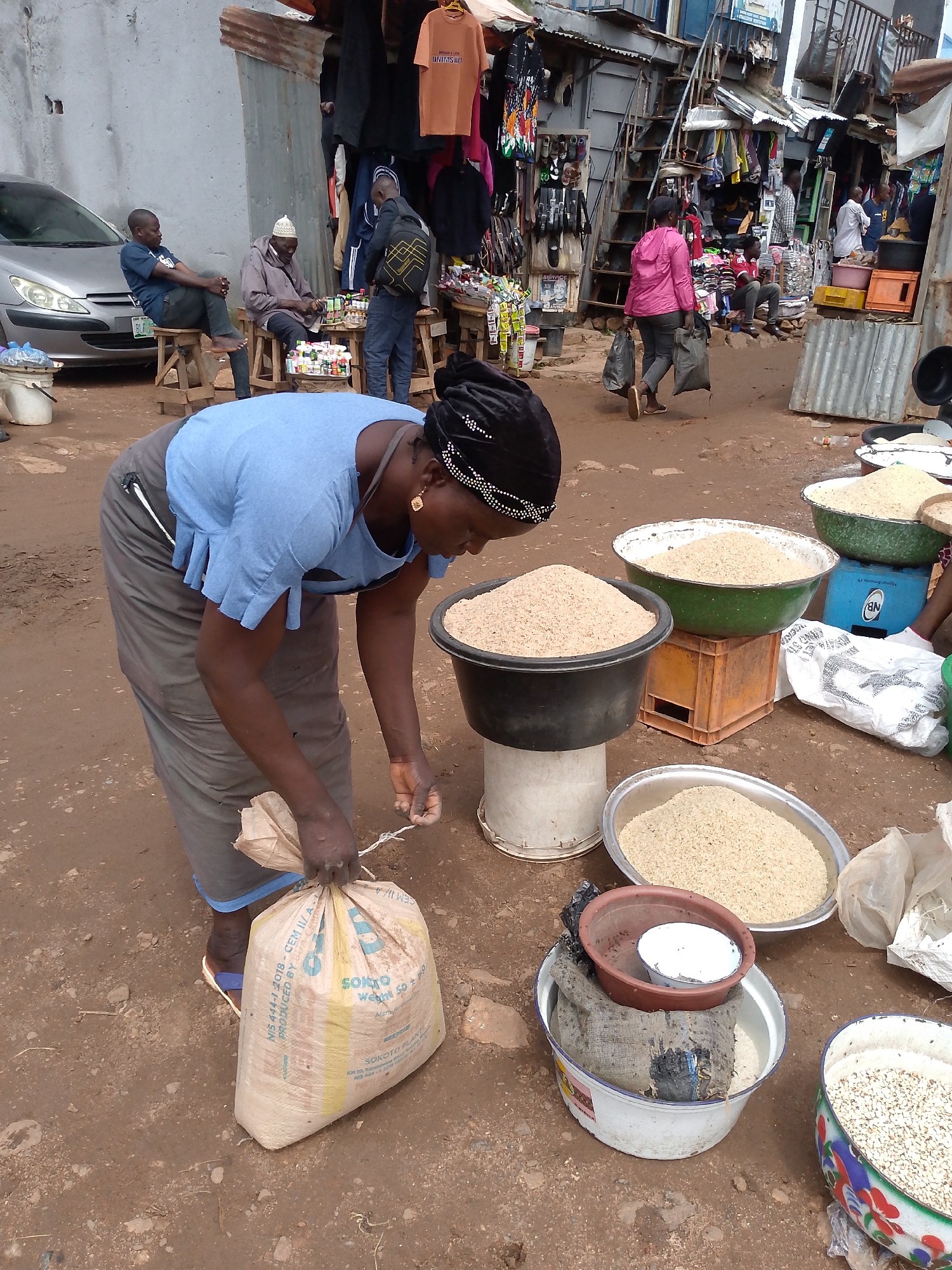 A Nigerian woman in a blue top and gray skirt is tying a bag of grain in a bustling outdoor market. She is bending over, her right hand holding a twine, and her left hand supporting the bag. The bag, made of woven material, has some text printed on it. In front of her, on the dusty ground, are several large bowls filled with grains. One large black bowl on a white pedestal contains a light brown grain, and to its right, a green bowl on an orange crate also holds a similar grain. Further to the right, a silver bowl is filled with lighter colored grains, and a colorful patterned bowl contains small white grains. Behind her, other market stalls are visible, displaying clothing and various goods. In the background, several people are scattered around the market, with a silver car parked to the left and a woman in a pink jacket carrying two black plastic bags walking away from the scene. The ground is unpaved and appears to be dirt. The overall atmosphere suggests a typical day in a Nigerian marketplace.