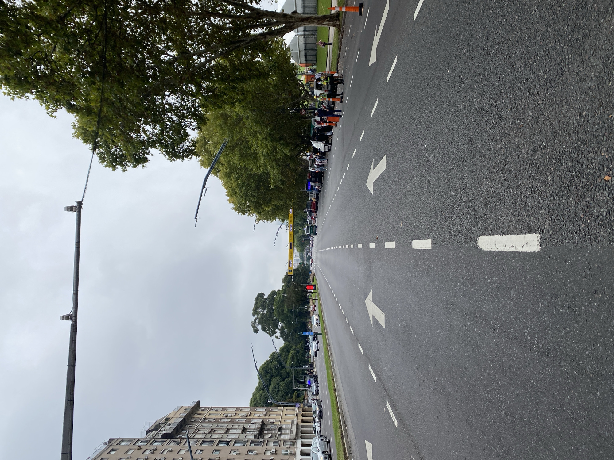 The image shows a street view in Buenos Aires, Argentina. A wide asphalt road stretches into the distance, marked with directional arrows and dashed white lines. The street is lined with trees on the left side, partially obscuring a row of parked cars and a multi-story building. People are gathered on the left side of the road, some wearing orange safety vests. Streetlights are visible along the left edge. The sky is overcast. An orange cone is visible on the edge of the road.
