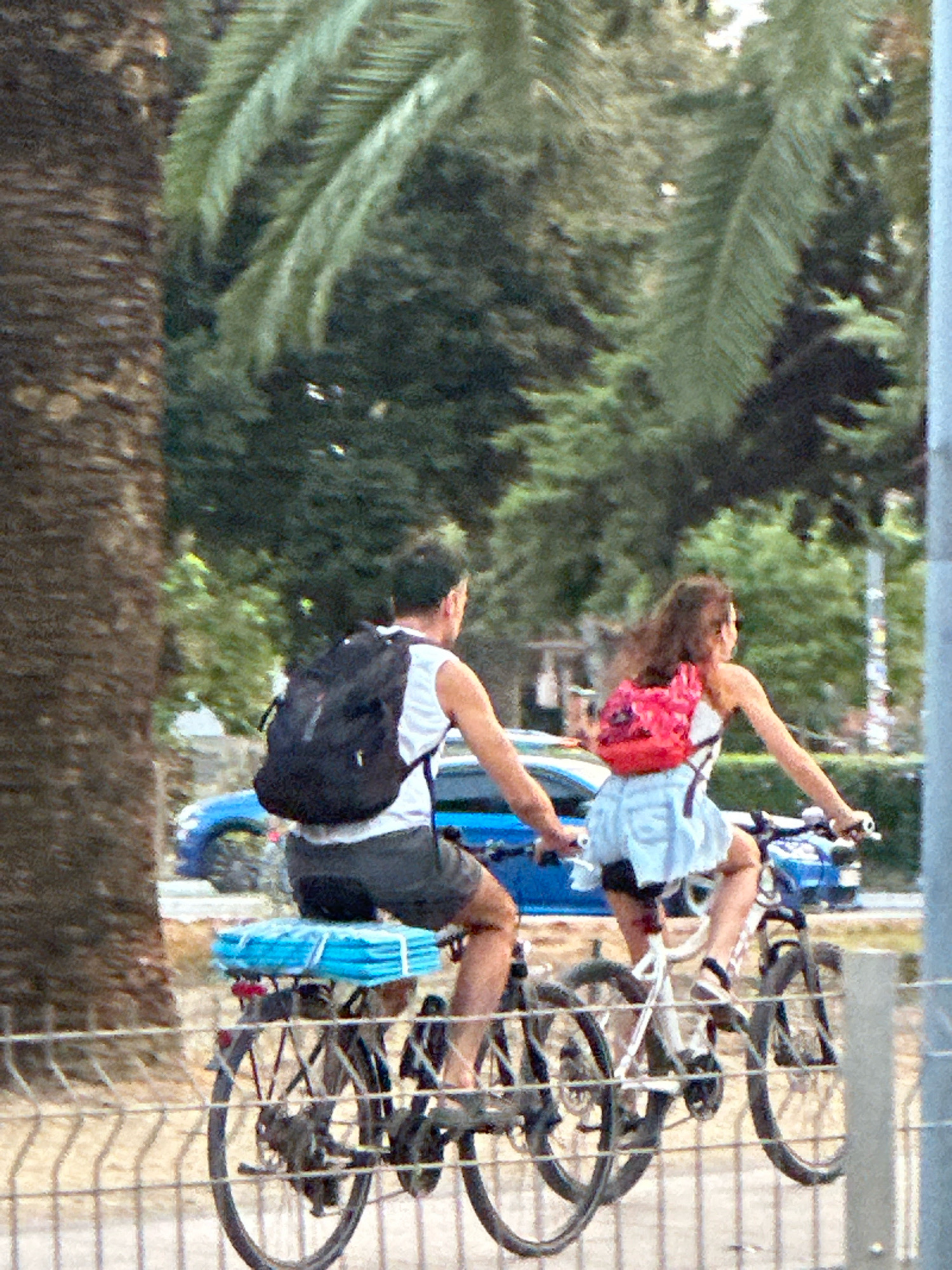 Two individuals are cycling on bicycles in Budva, Montenegro. The person in the foreground, viewed from the rear, is wearing a white tank top and dark shorts, with a black backpack. They are riding a bicycle with a blue rack holding a blue folded item. The second person, also viewed from the rear, is wearing a white top and a light-colored skirt, with a red backpack. This person is riding a white bicycle. Both bicycles are in motion. The background features lush green trees, including a palm tree on the left, and a blue vehicle. The scene appears to be a street or a path within a park-like setting, with a fence in the foreground.