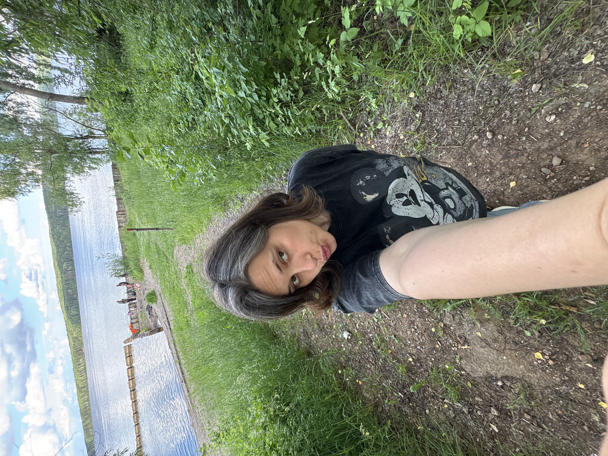 This is a selfie taken by a person with shoulder-length brown hair with streaks of gray. They are wearing a black t-shirt with a graphic design featuring skull-like imagery. The person is looking at the camera with a slightly curious or playful expression.
The scene appears to be outdoors, with lush green grass and vegetation in the foreground and background. To the left of the frame, a body of water, likely a lake or river, is visible, reflecting a cloudy sky. There is a wooden structure extending into the water, resembling a dock or pier, with a few people and what appear to be orange life vests on it. The ground is a mix of dirt and grass, with small pebbles scattered around. The overall atmosphere suggests a casual, outdoor activity, possibly a day by the water. The lighting indicates it is daytime. The location context provided is Chaikovsky, Russia.