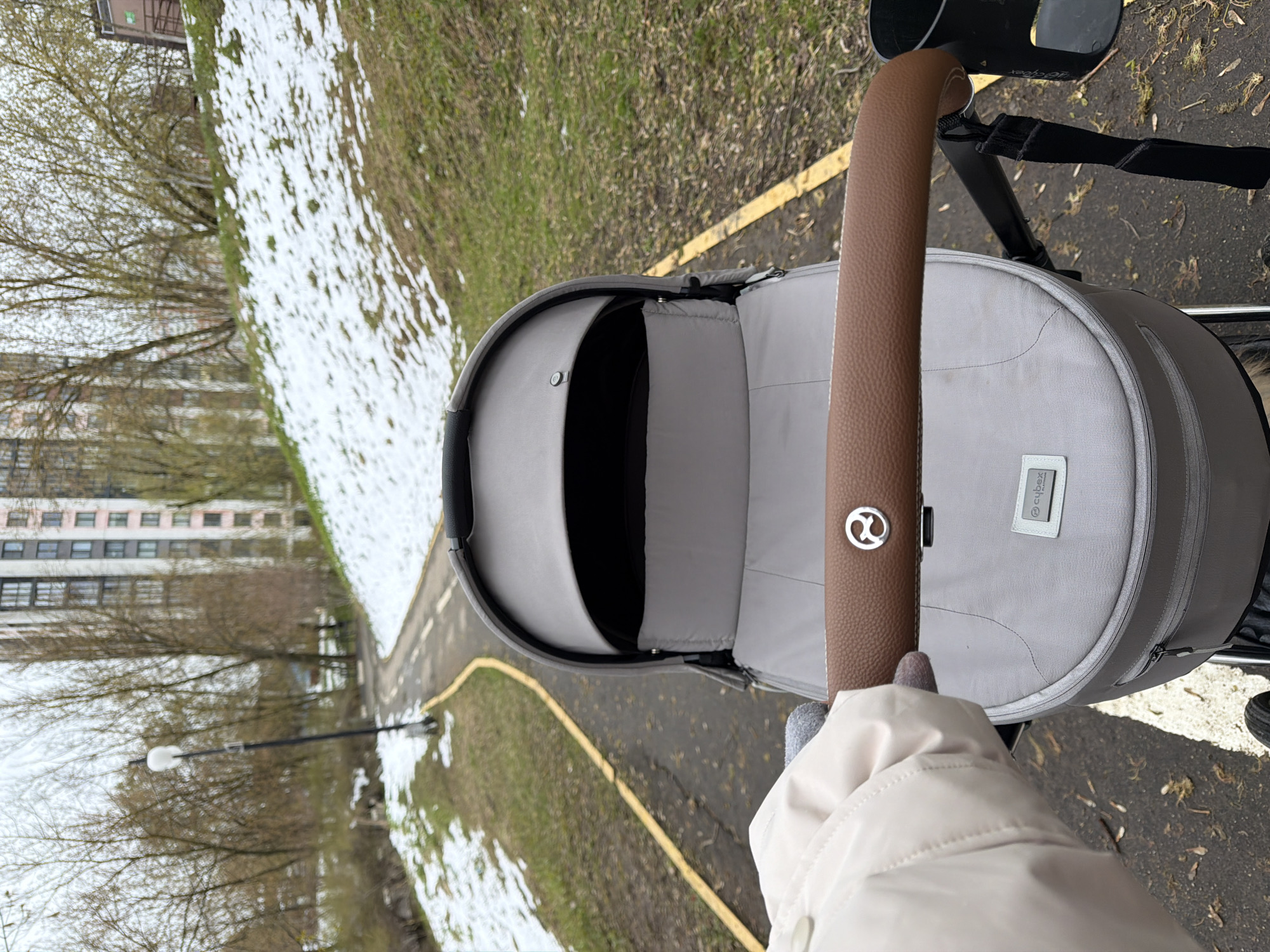 The image captures a first-person view of a person pushing a baby stroller outdoors in Khimki, Russia. The individual's hand, wearing a light-colored jacket or glove, holds the stroller's brown, textured handlebar, which features a silver Cybex logo. The stroller itself is light grey with a dark interior lining, and a small "Cybex" tag is visible on its side.
The setting is an outdoor paved path with a distinct yellow line, flanked by grassy areas. Patches of melting snow are scattered across the grass, suggesting a late winter or early spring season. Bare trees stand in the background, further indicating the time of year. In the distance, a multi-story residential building with numerous windows is visible, alongside a street lamp. The lighting appears soft and overcast, typical of daytime conditions. The primary activity depicted is a casual stroll with a baby carriage.