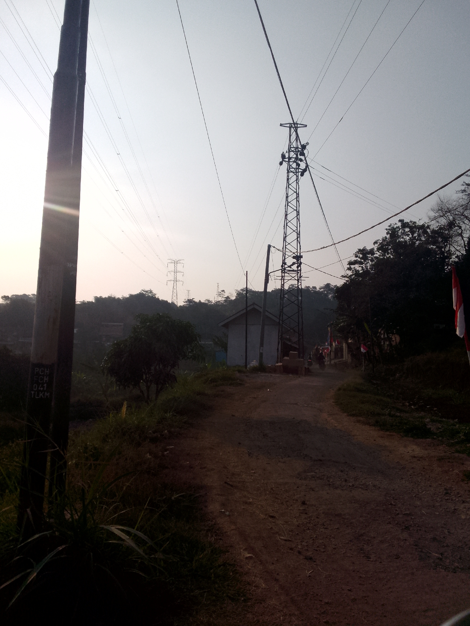 The image depicts a dirt road in a rural or semi-rural area, likely in Padalarang, Indonesia, given the presence of Indonesian flags. The scene is dominated by a large power line tower and a utility pole on the left. The sky is overcast and appears to be early morning or late afternoon, indicated by the soft light and shadows. The road is unpaved and shows signs of wear, with scattered rocks and dust.
On the left side of the road, a tall, dark utility pole stands prominently. It has some markings visible on its surface, including the text "PCH," "FCH," "041," and "TLKM." A thin patch of tall grass grows at the base of this pole. The sun is creating a lens flare effect near the top left of the image, shining from behind the utility pole.
Further down the road, a tall, lattice-style power line tower is visible. It supports multiple wires that stretch across the frame. Small structures, possibly houses, are scattered on the hilly landscape behind the road and the power line tower. Some trees with dark foliage are present on the right side of the road, obscuring parts of the background.
Several Indonesian flags, red and white, are visible on poles along the right side of the road. Two motorcycles and possibly a few people are present further down the road, moving away from the camera. One of the motorcycles appears to be kicking up dust as it travels. The overall impression is a tranquil, everyday scene in a developing area, with infrastructure for electricity and signs of local life.