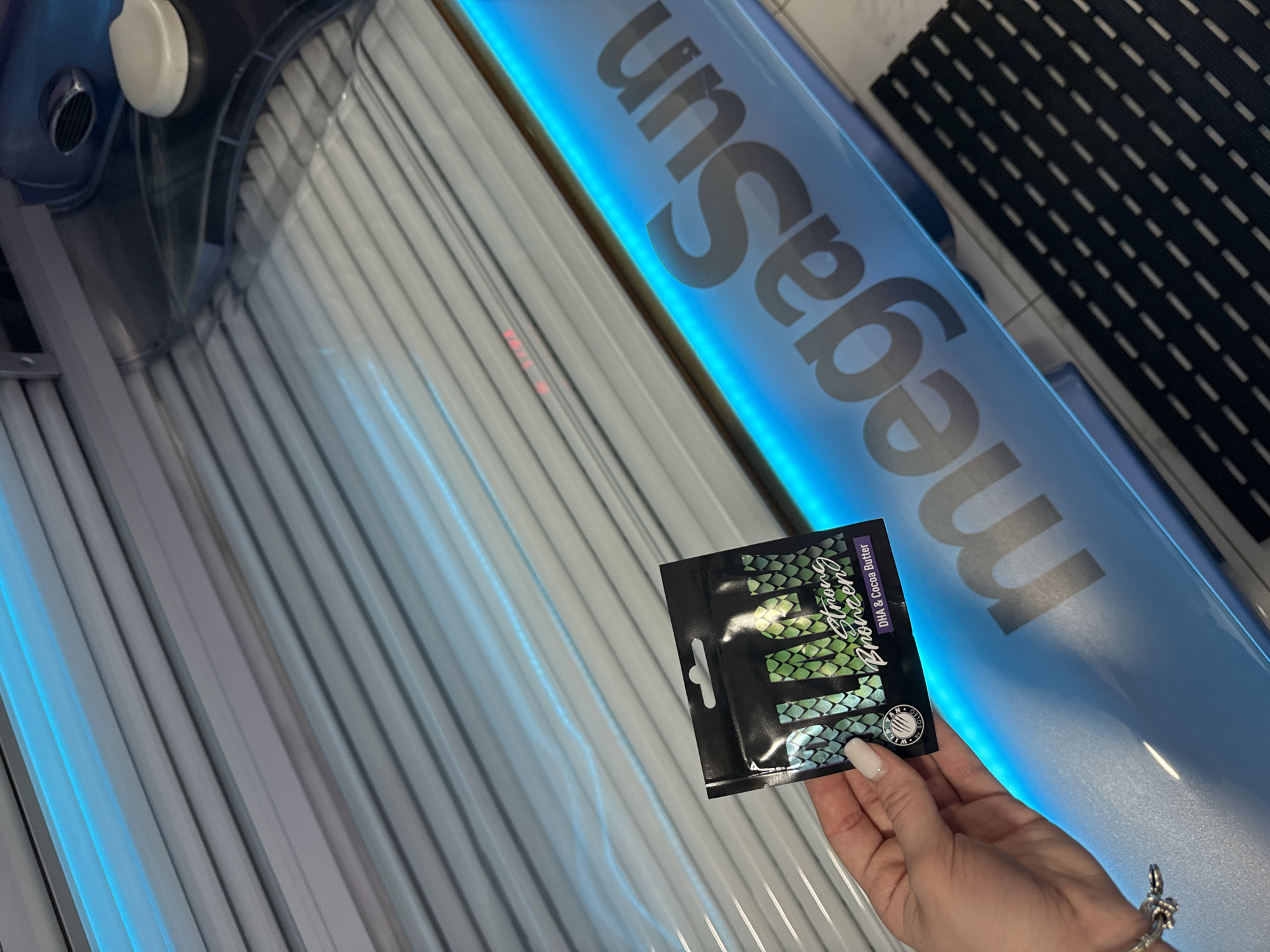 A tanning bed, labeled "megaSun", is visible. A hand with manicured nails holds a black packet labeled "Strong Bronzer" with "DHA & Cocoa Butter". The tanning bed features rows of UV lamps. The location is impossible to ascertain based on visual information alone. No people are visible except for the hand. The scene appears to be indoors.