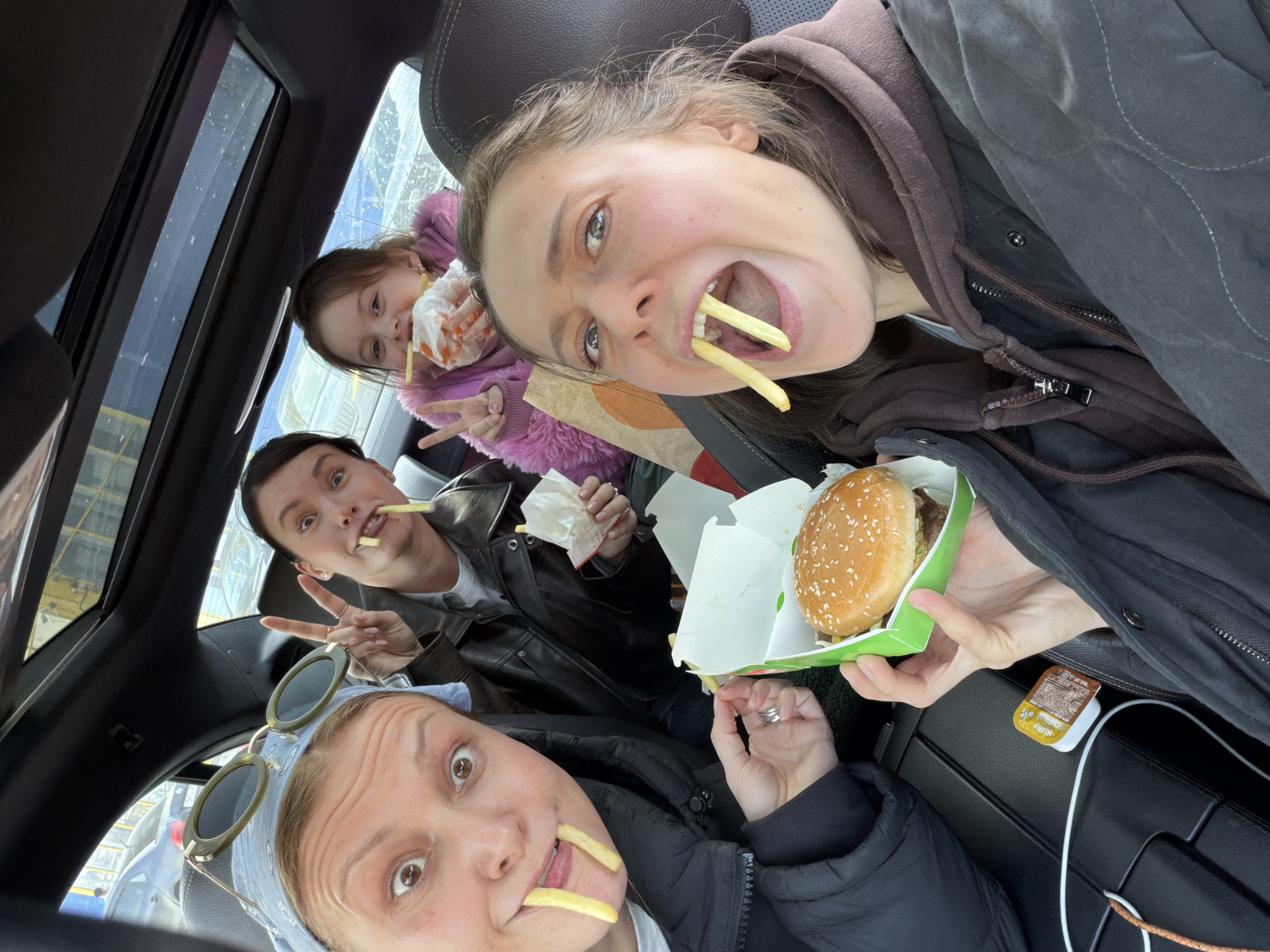 Four individuals, three adult females and one female child, are depicted inside a dark-colored vehicle in Khimki, Russia, engaging with fast food. The scene is captured from a slightly elevated perspective, presumably from one of the occupants.
In the front-left seat, an adult female with light-colored hair, wearing a light blue bandana and olive-green sunglasses pushed onto her head, looks directly at the camera with wide eyes. She is wearing a dark puffer jacket over a light top. Two french fries are intentionally placed to protrude from her open mouth. Her left hand holds a green and white cardboard box containing a sesame seed bun hamburger. A silver ring is visible on her left ring finger.
Behind her, in the rear-left seat, another adult female with dark hair, wearing a black leather jacket, also looks towards the camera with wide eyes. She makes a "peace" sign with her right hand. A single french fry protrudes from her mouth. Her left hand holds crumpled white paper, consistent with a fast-food fry wrapper.
In the rear-right seat, a young female child with brown hair, dressed in a pink fluffy jacket, looks towards the camera. A french fry protrudes from her mouth, and she holds a white paper bag with orange markings, containing what appears to be more french fries. She wears small earrings.
In the front-right seat, another adult female with brown hair, wearing a dark grey quilted jacket over a brown hooded sweatshirt, stares directly at the camera with wide eyes and a wide-open mouth. Two french fries are inserted into her mouth. Her left hand is partially visible near a fast-food container.
The vehicle interior features dark upholstery, likely leather, and a dark headliner. A sunroof is visible above, revealing a glimpse of the sky and a yellow structure in the distance. The center console between the front seats displays a small yellow Heinz sauce packet, bearing Russian text. Through the car windows, other vehicles are visible, suggesting a parking lot or drive-thru setting.